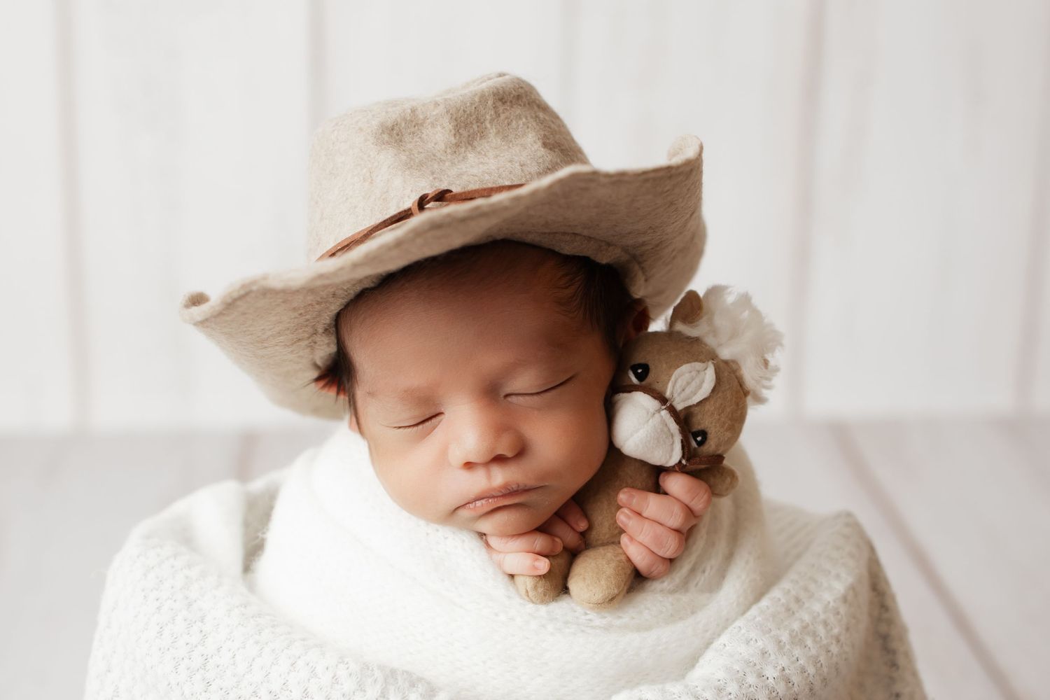 Newborn baby sleeping in cream wrap wearing cowboy hat while holding small plush toy deer.