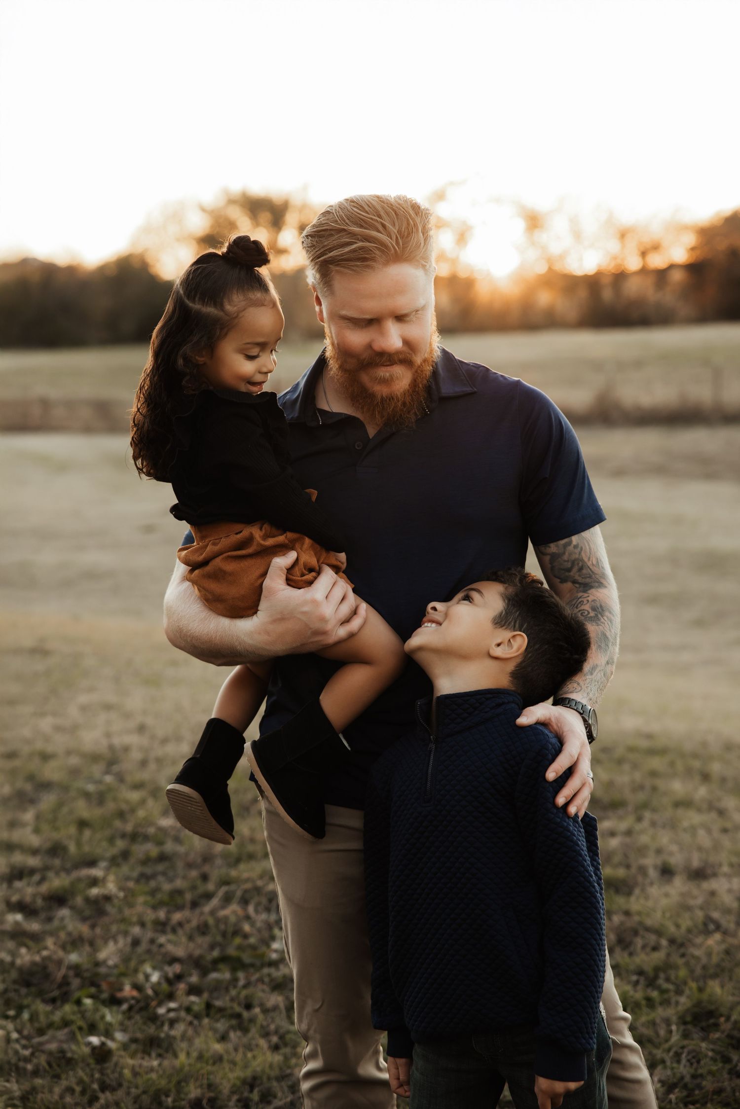A red-bearded tattooed father holds his young daughter while embracing his son in a field.