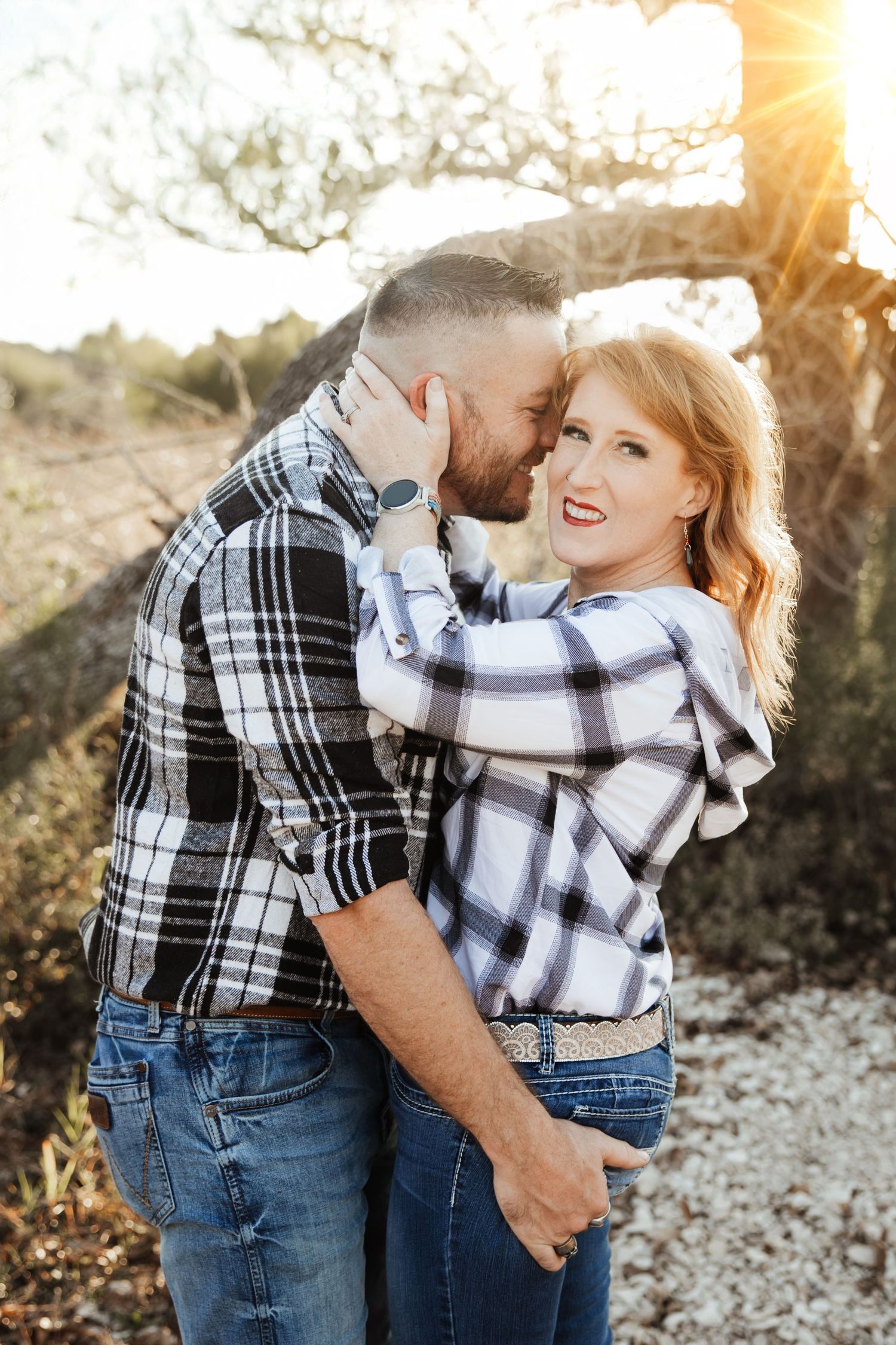 A couple in matching plaid shirts embrace romantically under a sunlit tree outdoors.