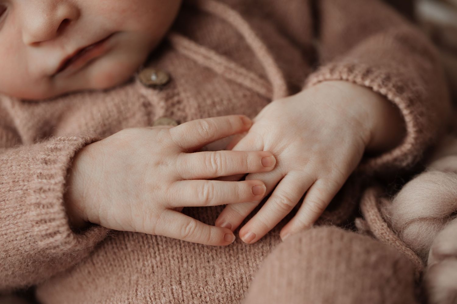 Close-up of a sleeping newborn baby's tiny hands in a soft pink knit sweater.