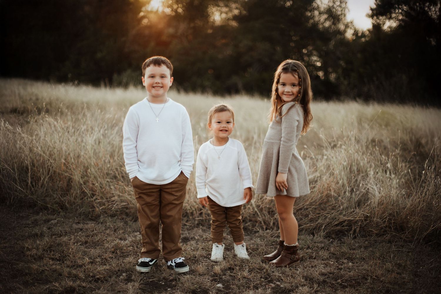 Three young siblings stand together smiling in a sunlit golden field during an outdoor family photo session.