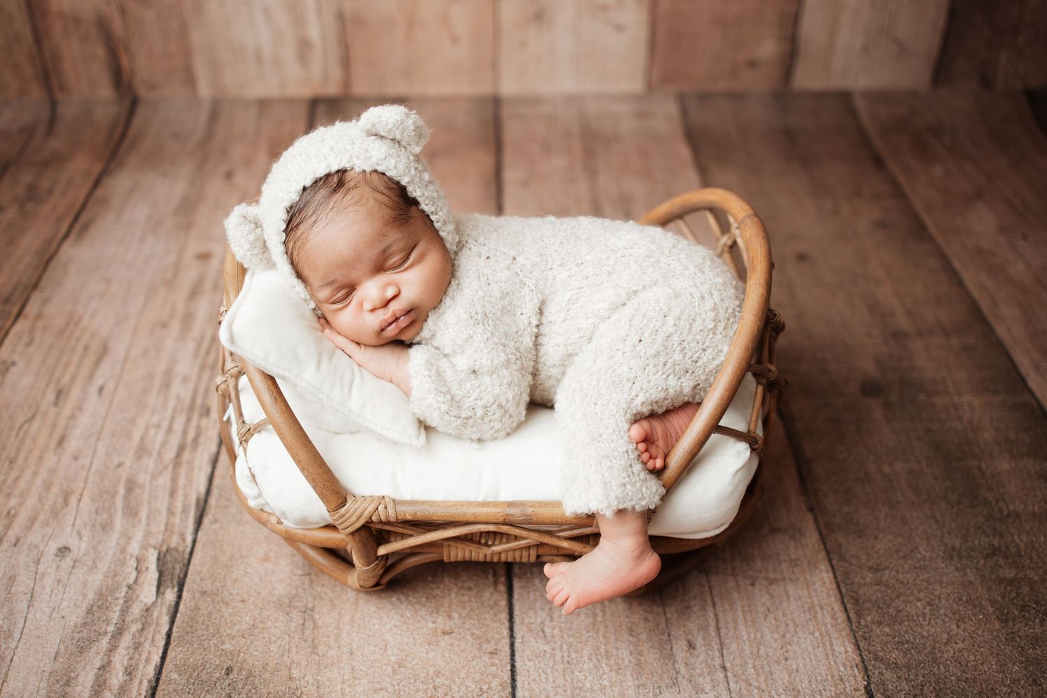 A newborn baby in a white fuzzy bear outfit sleeps peacefully in a small wicker basket.