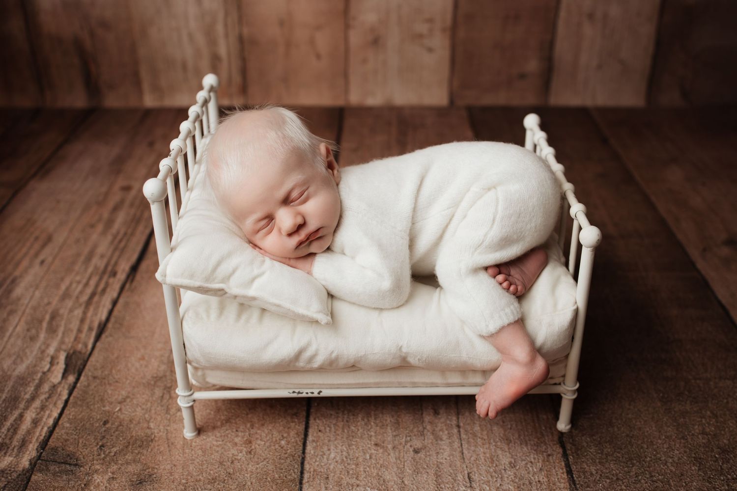 Sleeping newborn baby in white romper curled on tiny white metal bed with wood floor backdrop.