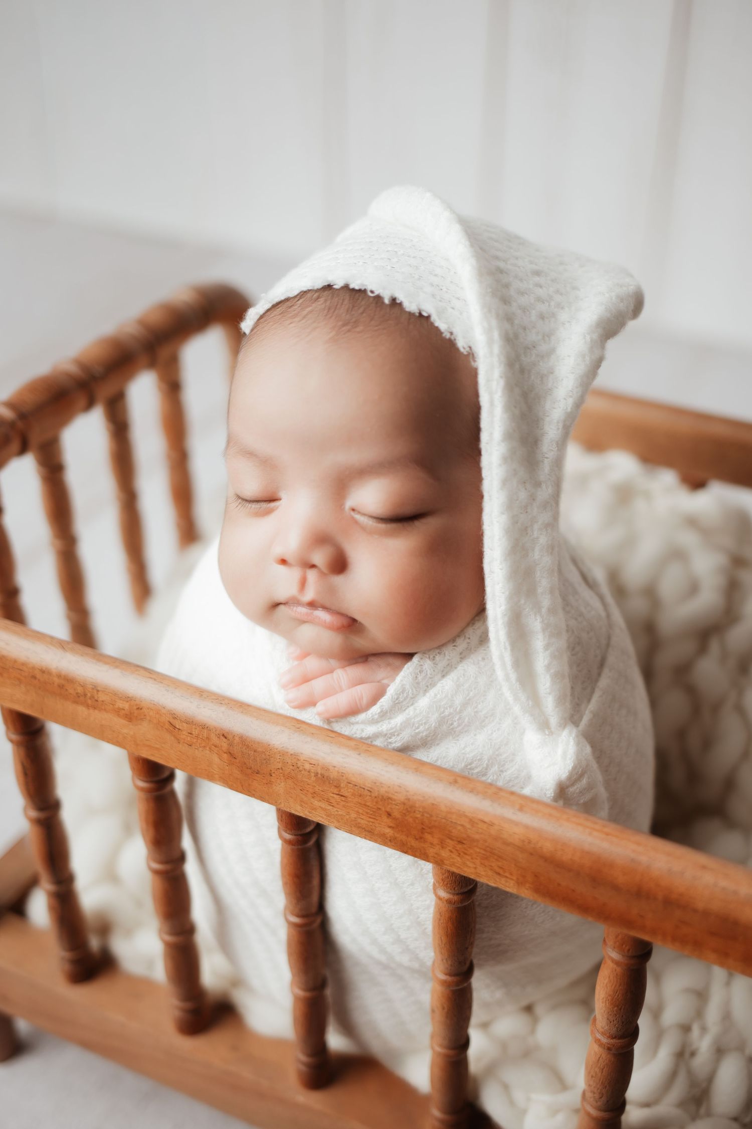 Newborn baby wrapped in white swaddle with bunny ear bonnet sleeping in wooden mini crib.