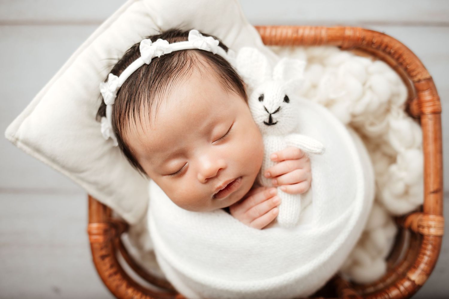 Sleeping newborn baby girl in a wicker basket holding a white knit bunny toy, wearing a floral headband.