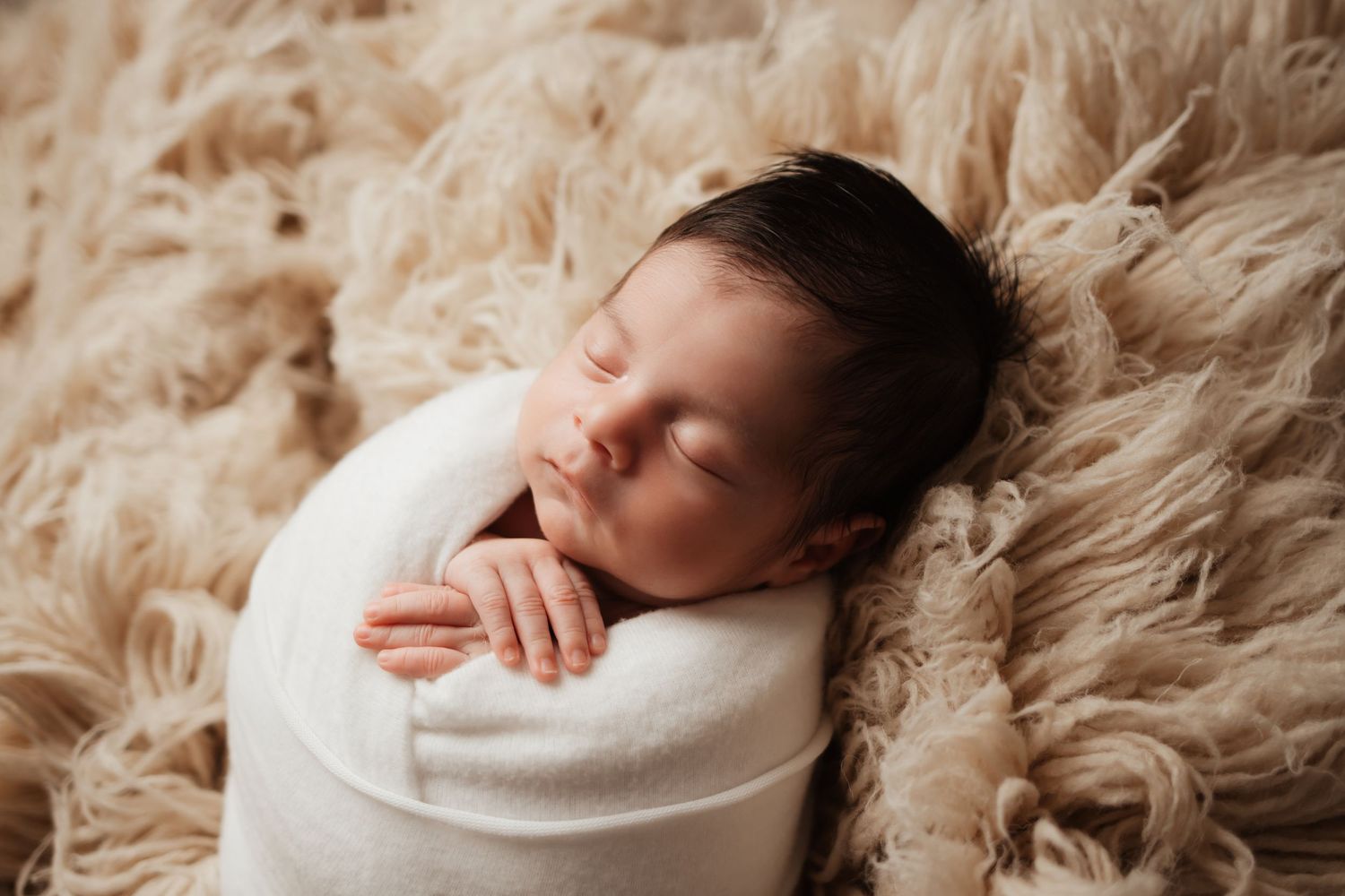 Sleeping newborn baby swaddled in white wrap on fluffy beige fur backdrop.