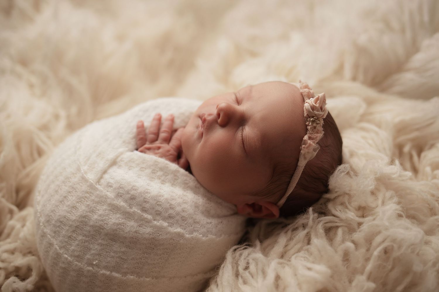 Sleeping newborn baby girl swaddled in white wrap with pink floral headband on fur.