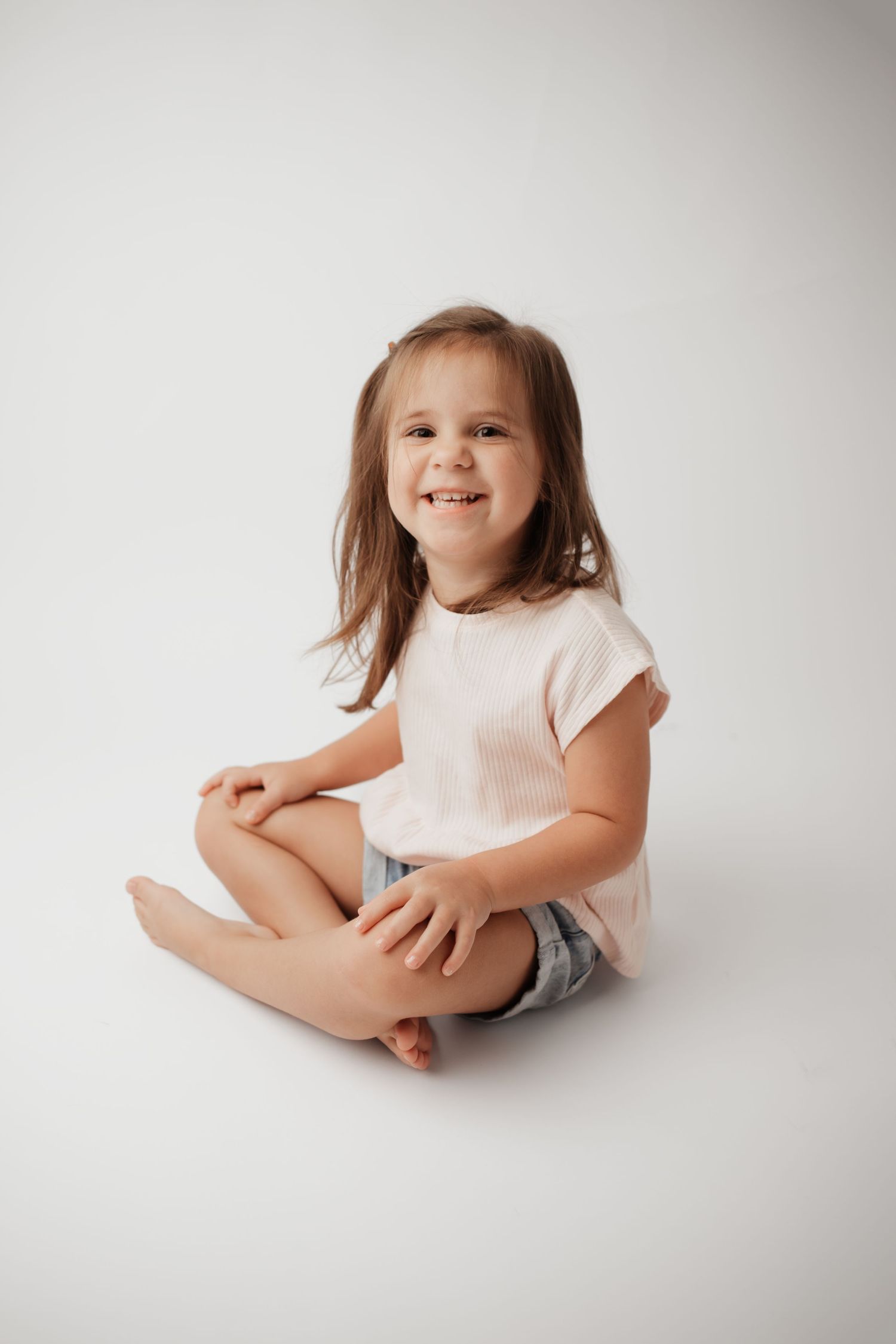 Happy little girl with brown hair in white shirt and jean shorts sitting on white background.