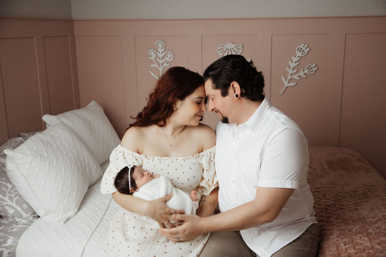 Loving new parents touching foreheads while holding swaddled newborn baby in pink nursery bedroom.