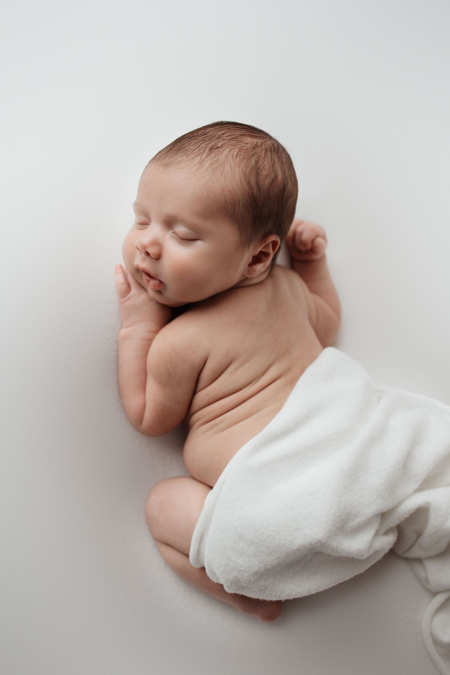 Peaceful sleeping newborn baby curled up on white blanket in a serene newborn photography pose.