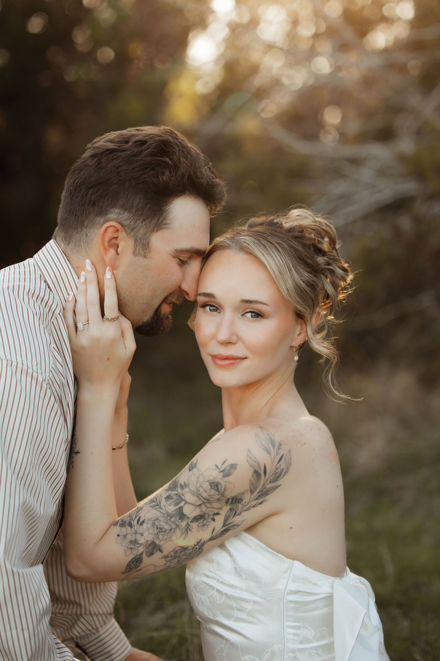 A bride with a floral arm tattoo smiles as her groom nuzzles her cheek during a golden hour outdoor wedding shoot.