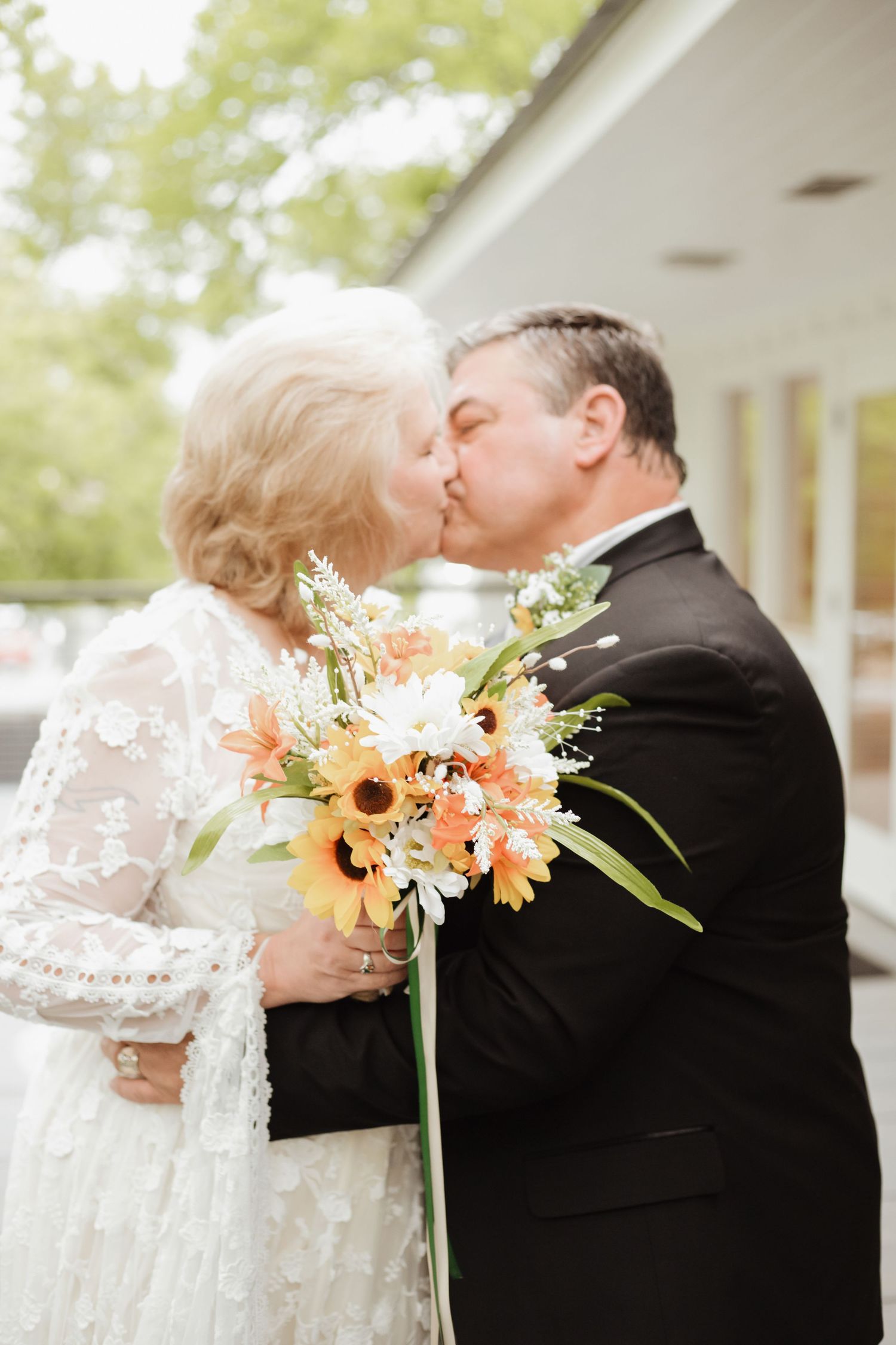 Older bride and groom sharing a kiss, bride holding sunflower bouquet, wearing lace dress and black tuxedo.