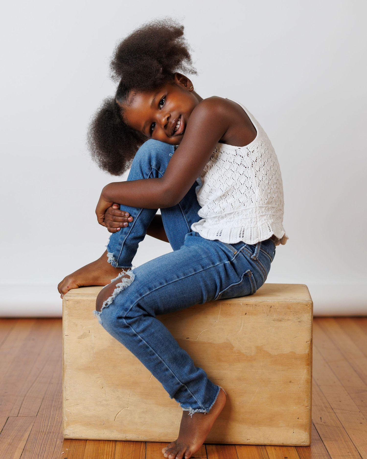 Young Black girl sitting on wooden box in white top and jeans, smiling at camera.