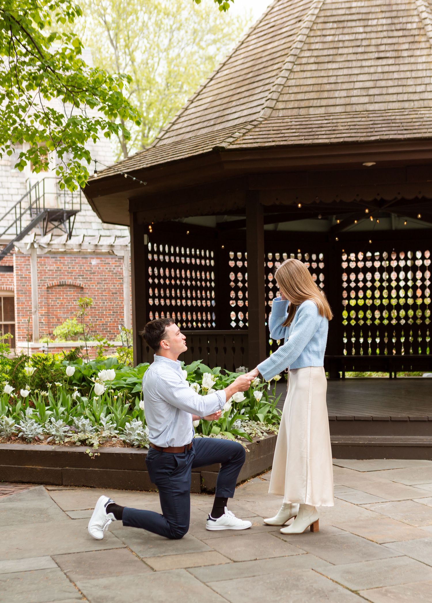Man kneeling on one knee proposing to woman near a gazebo in a lush garden setting.