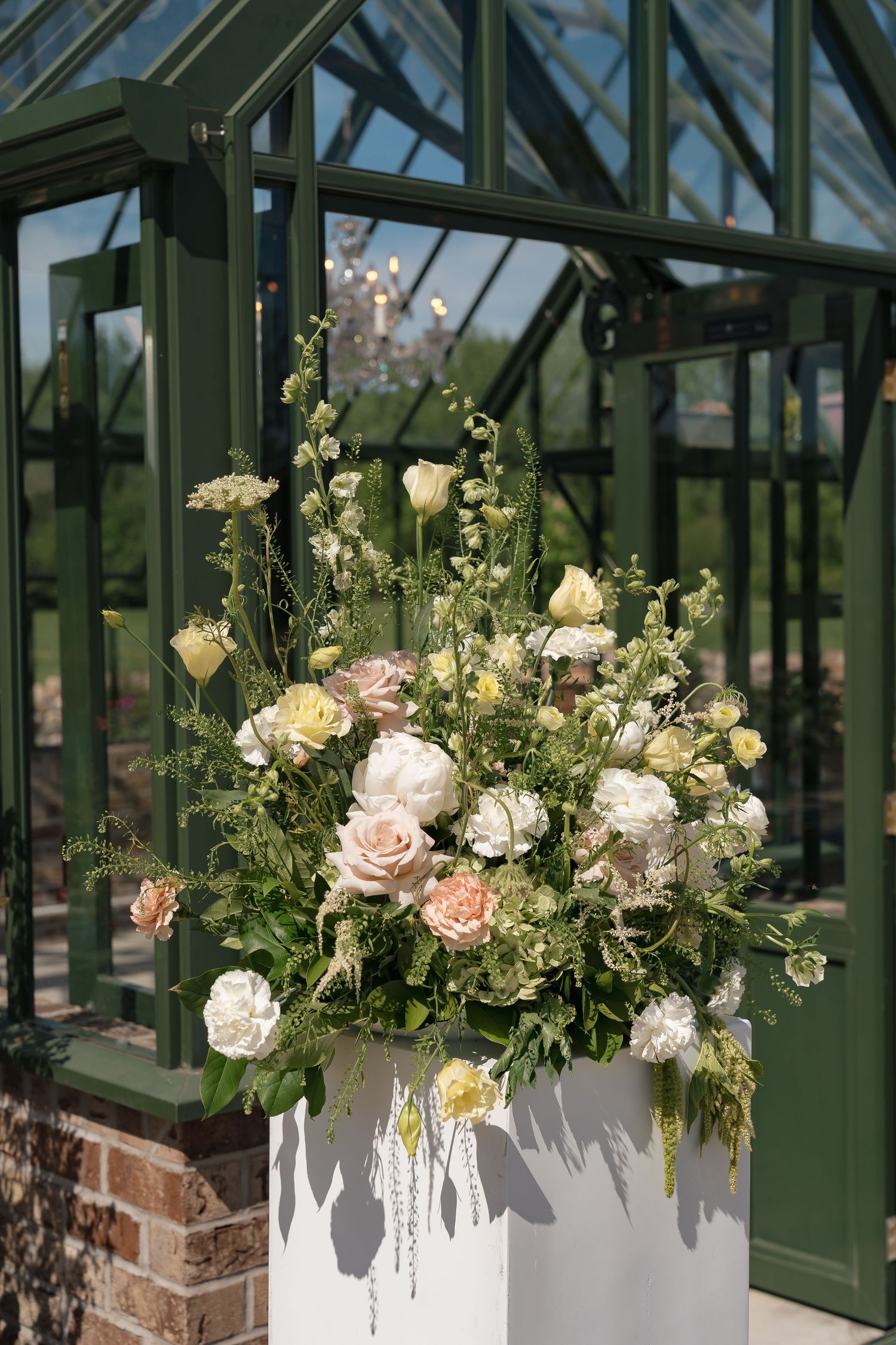 Elegant floral arrangement of blush roses, white peonies, and greenery in white vase outside a green greenhouse.