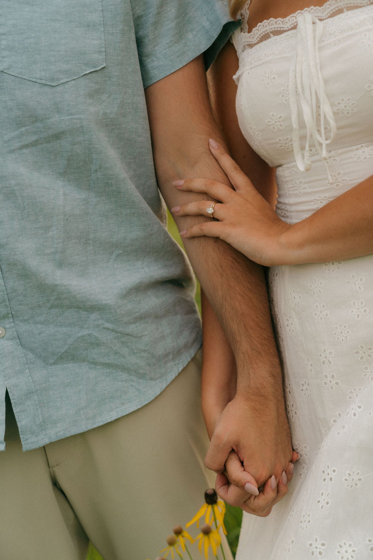 Couple holding hands outdoors, woman wearing engagement ring in white dress beside man in light blue shirt.