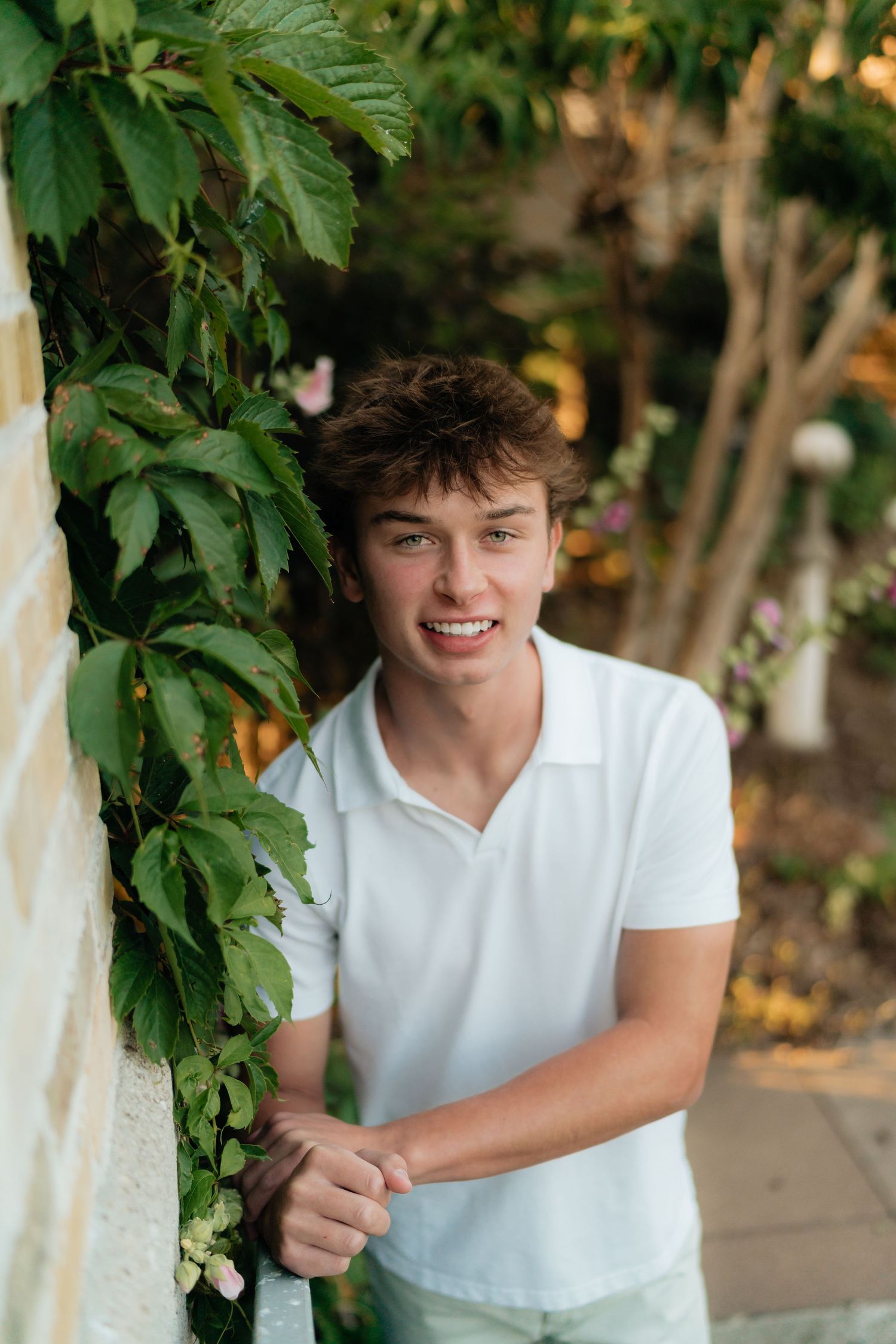 Smiling young man in white polo shirt leaning against ivy-covered wall in lush green garden setting.
