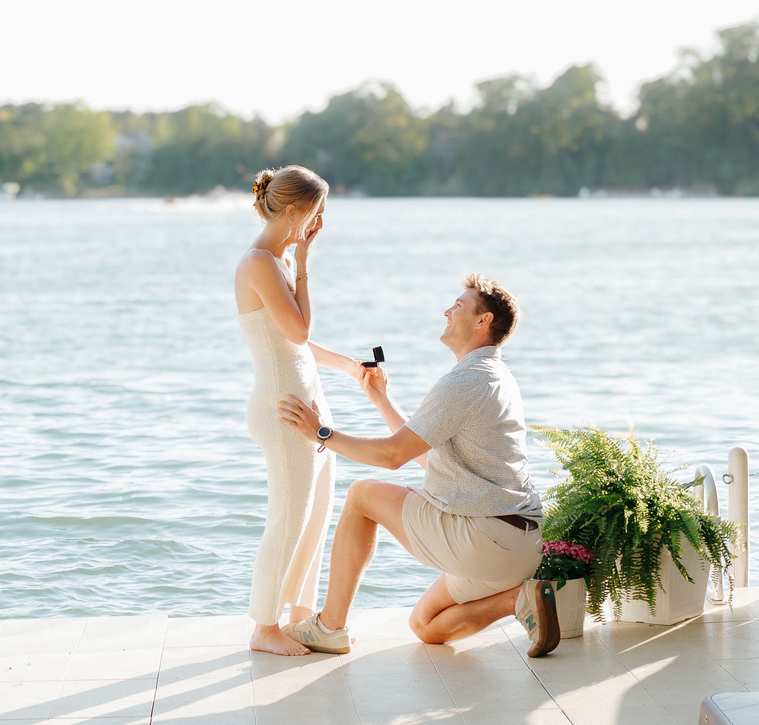A man is proposing to a woman on a dock by a lake with a bouquet of flowers.