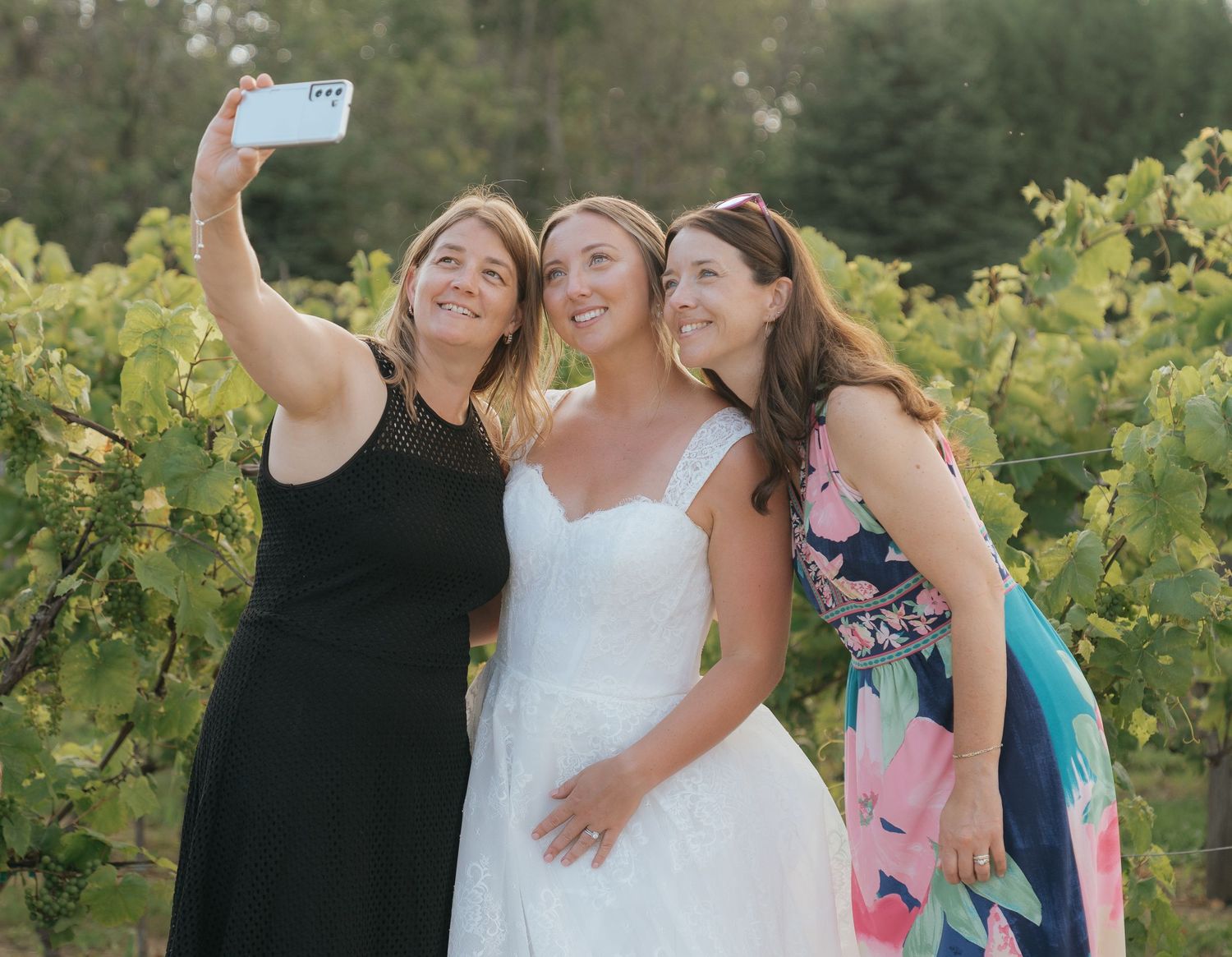 Three women in formal dresses taking a selfie together in a vineyard setting.