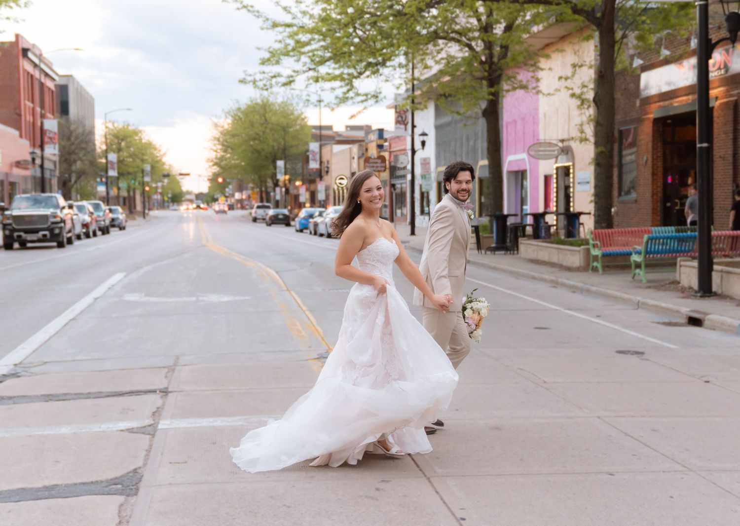 A woman in a flowing white wedding dress walks down a city street lined with shops and trees.