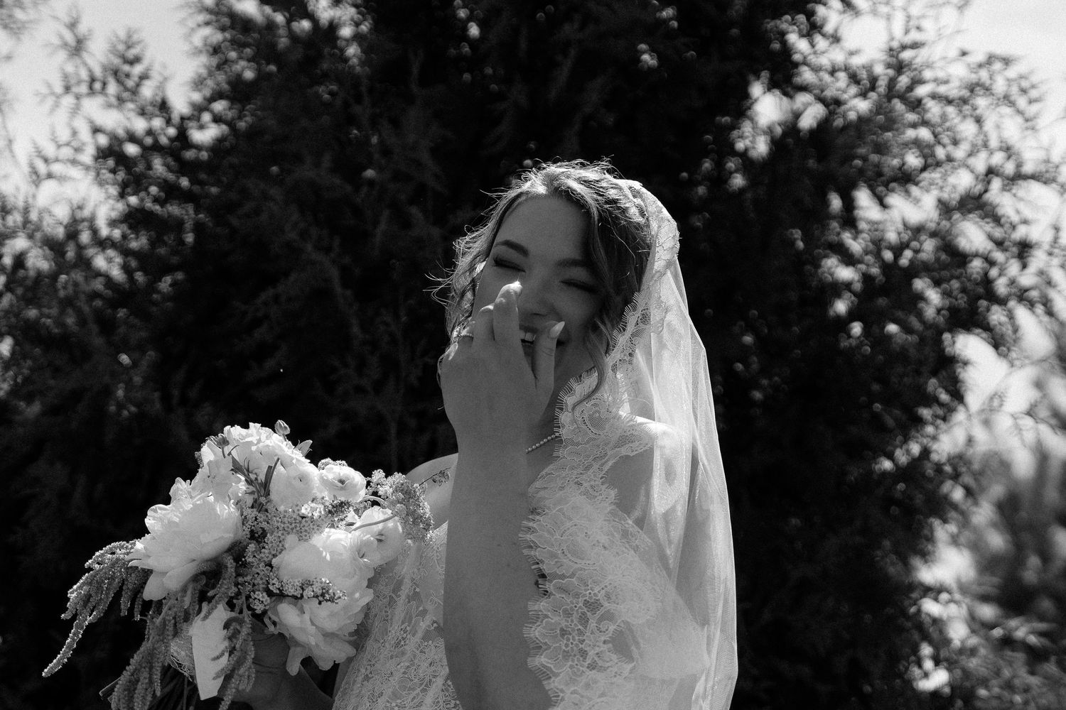 Black and white photo of a bride in lace gown and veil, holding bouquet, laughing outdoors near trees.