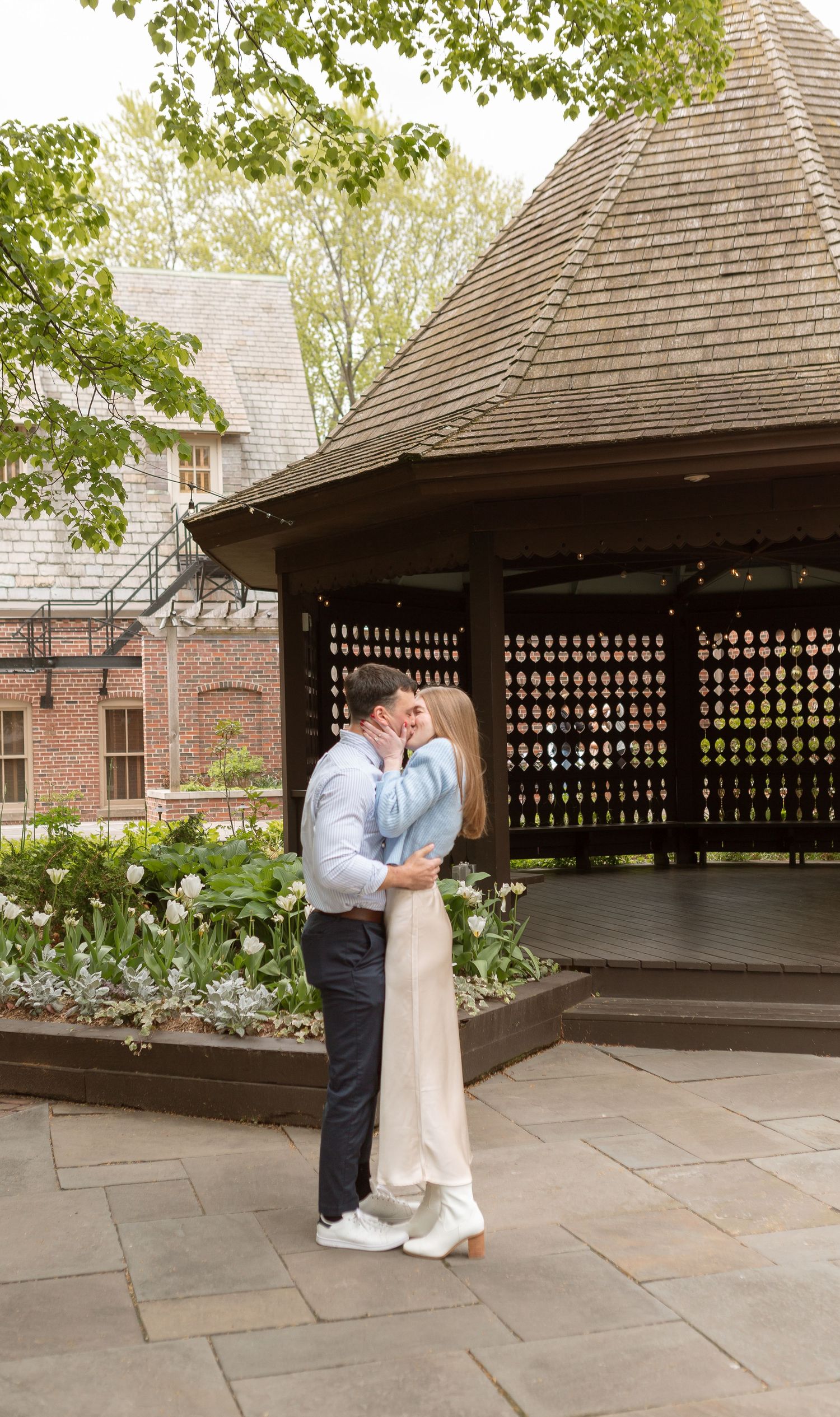 A couple embracing in a garden with a wooden gazebo in the background.