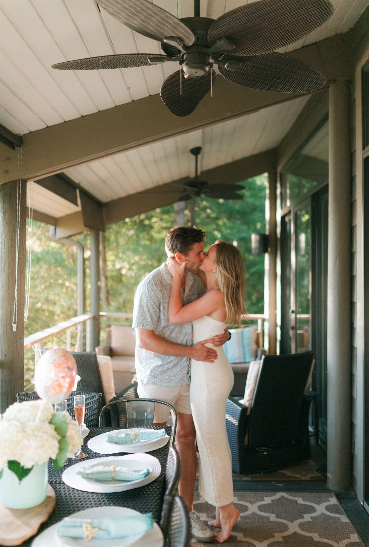 A couple embracing and kissing on a porch with a ceiling fan and outdoor furniture.