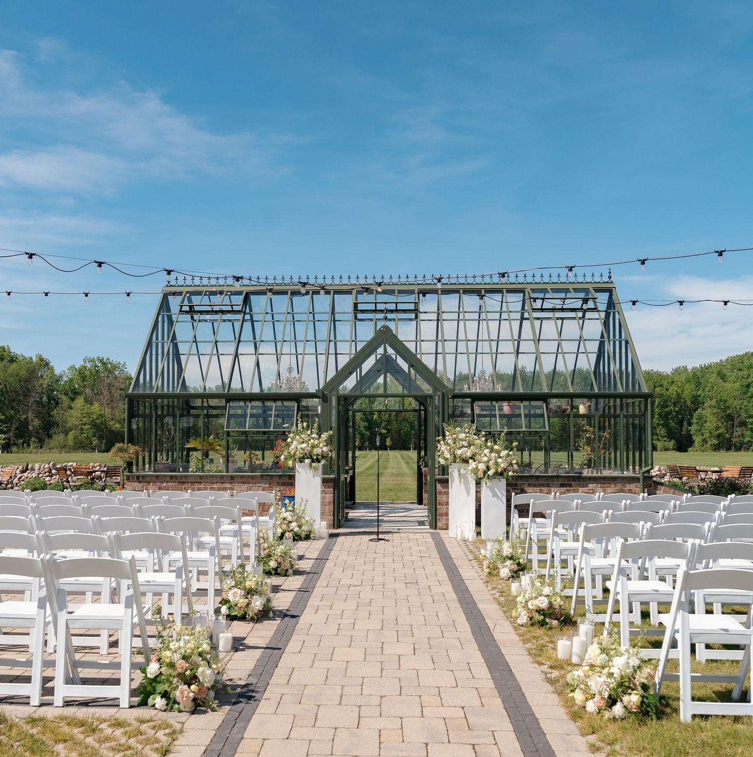 Outdoor wedding ceremony setup with white chairs flanking a brick aisle leading to a glass greenhouse venue.