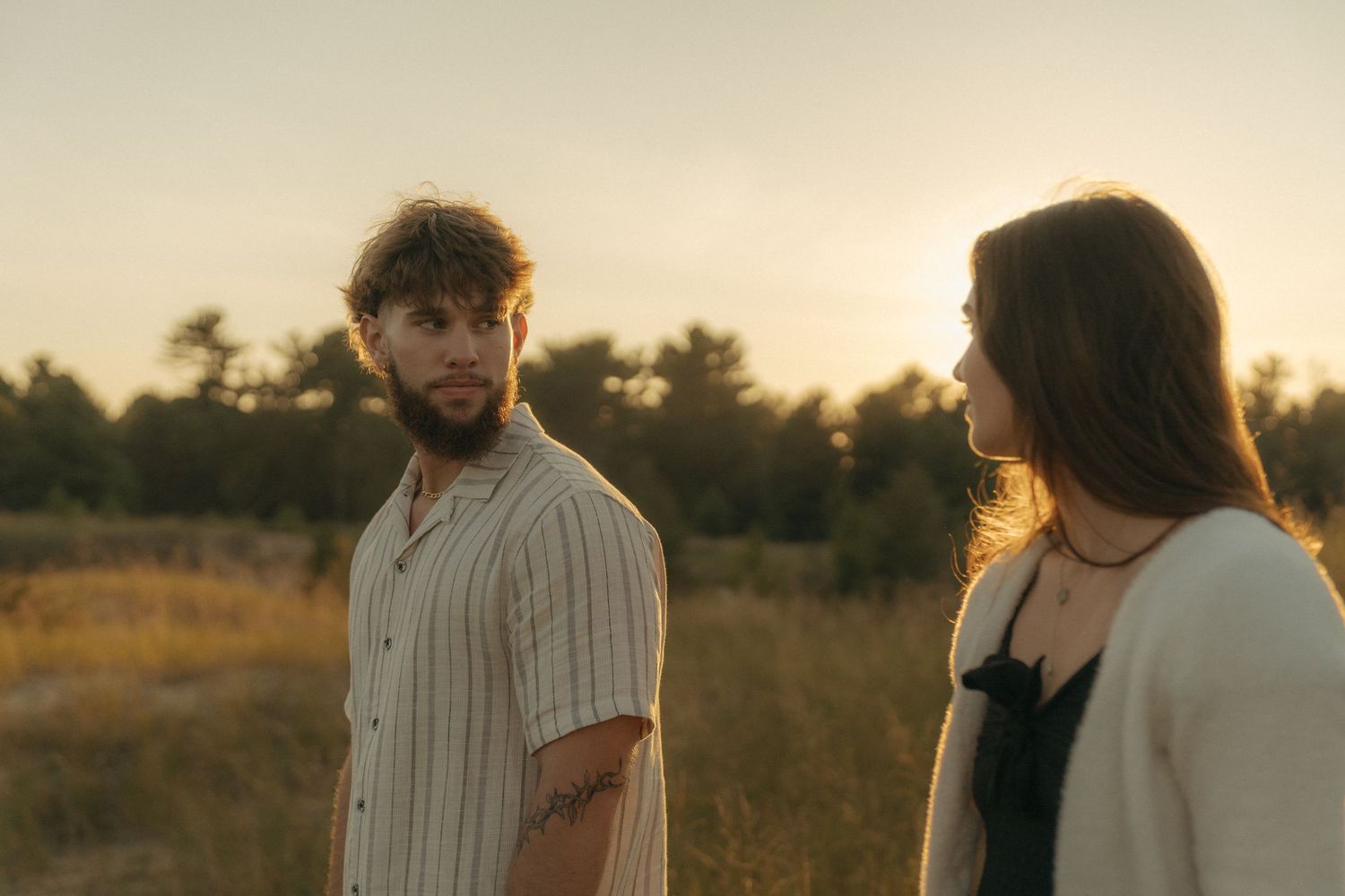 Two people, a man and a woman, standing in a field at sunset, looking at each other.