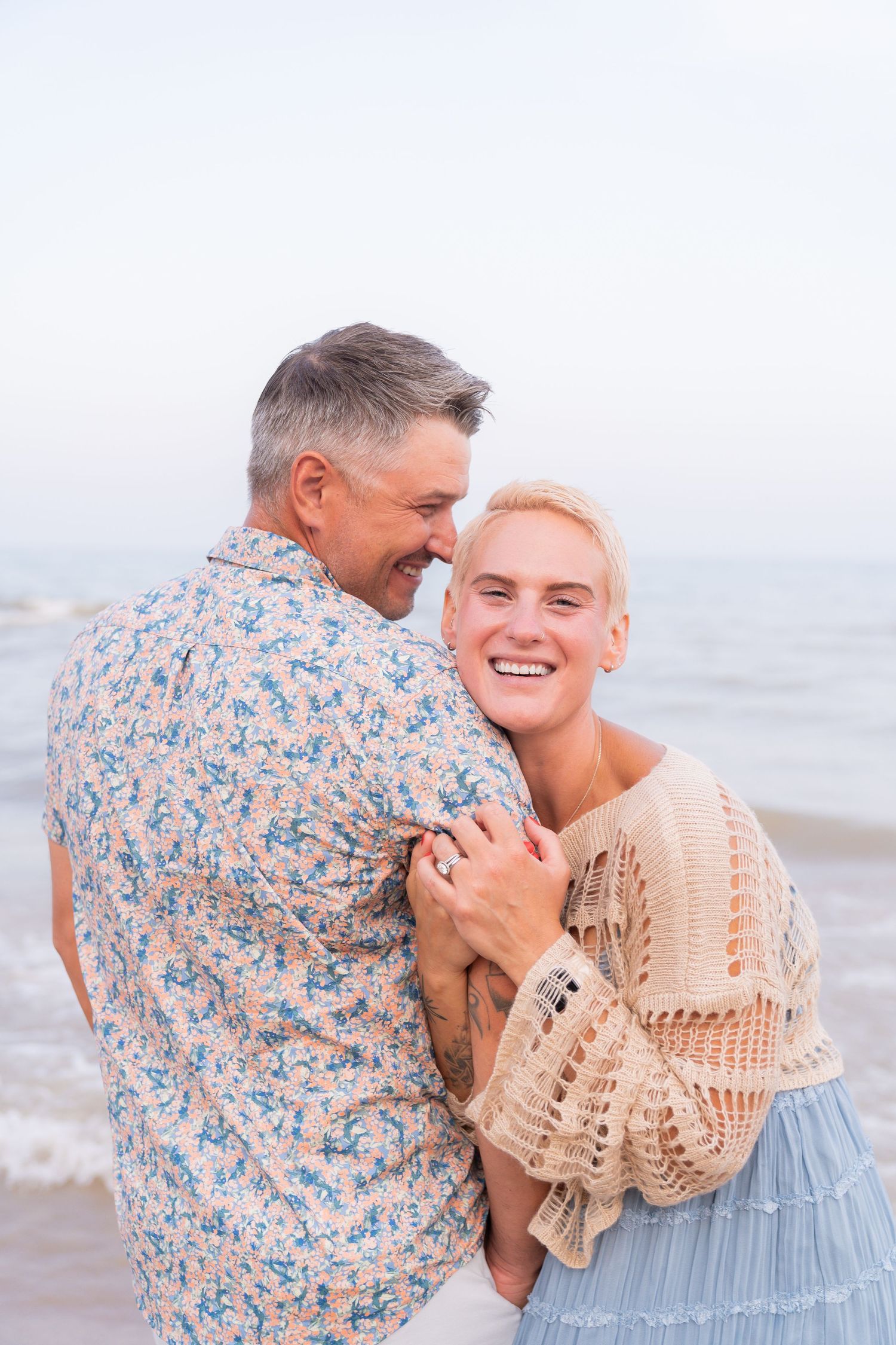An affectionate couple embracing on a beach, with the man wearing a floral shirt and the woman wearing a beige sweater.