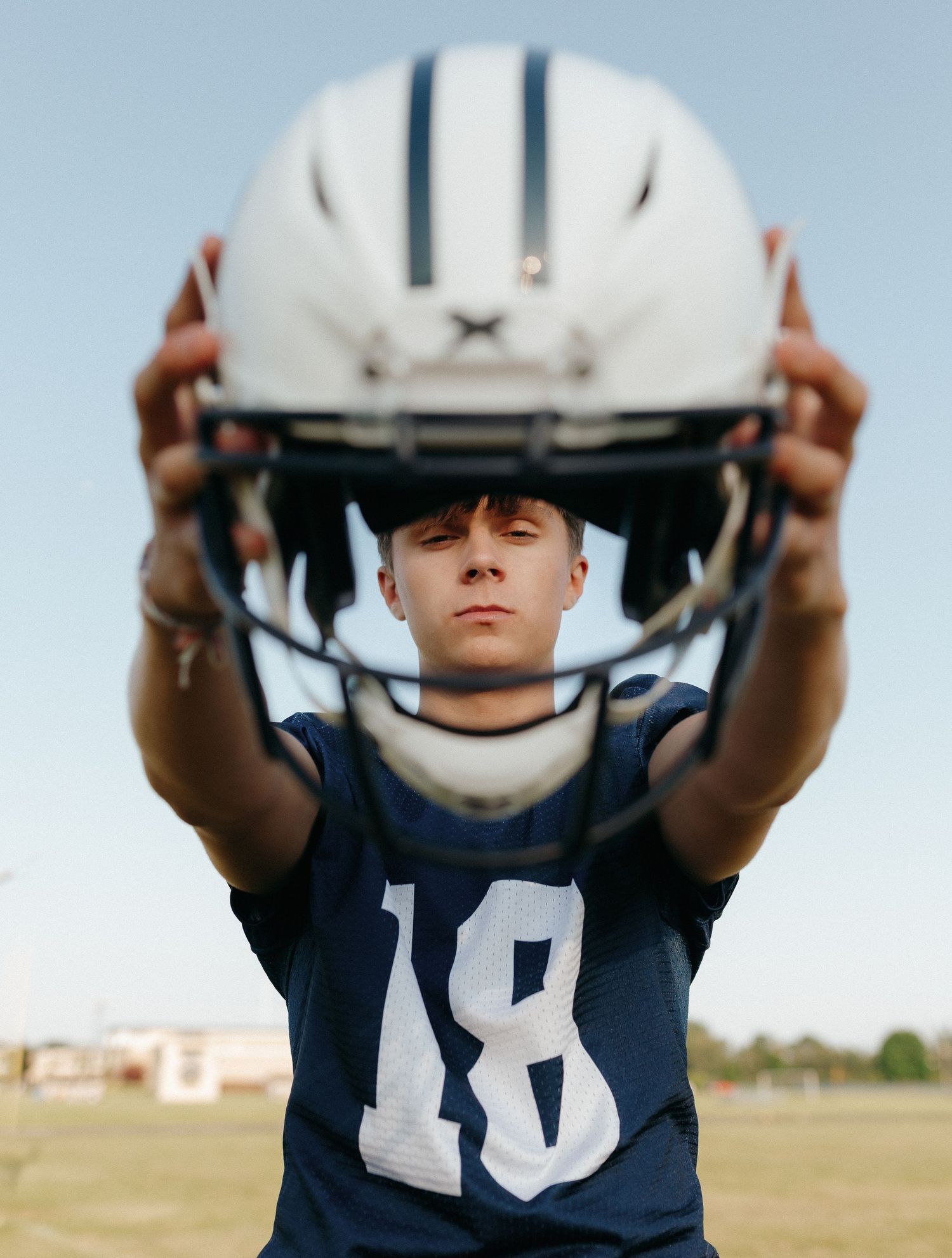 A young football player wearing a helmet and jersey stands on a field, holding the helmet in front of his face.