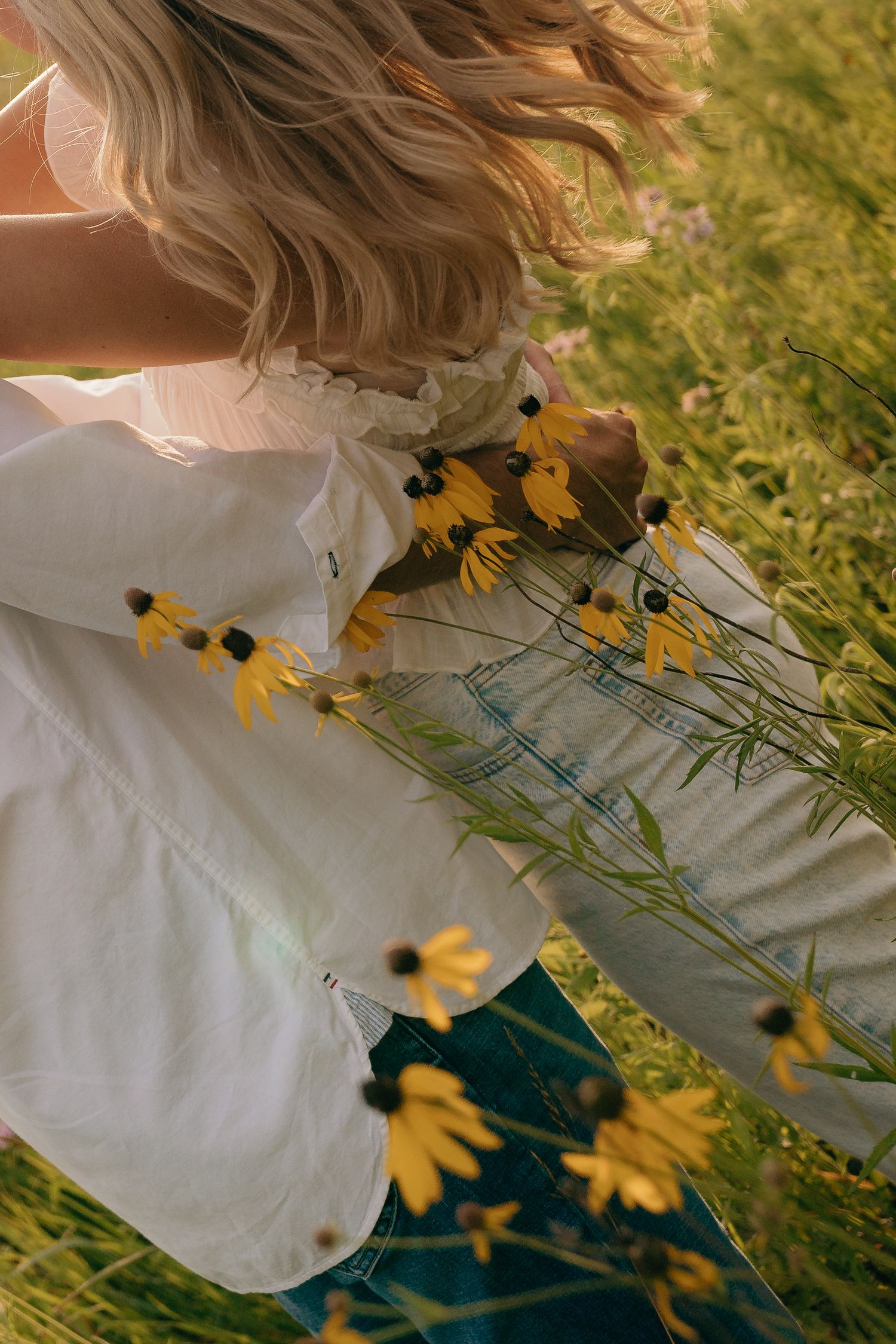 Blonde woman in white blouse holding yellow black-eyed Susan wildflowers in a lush green meadow field.