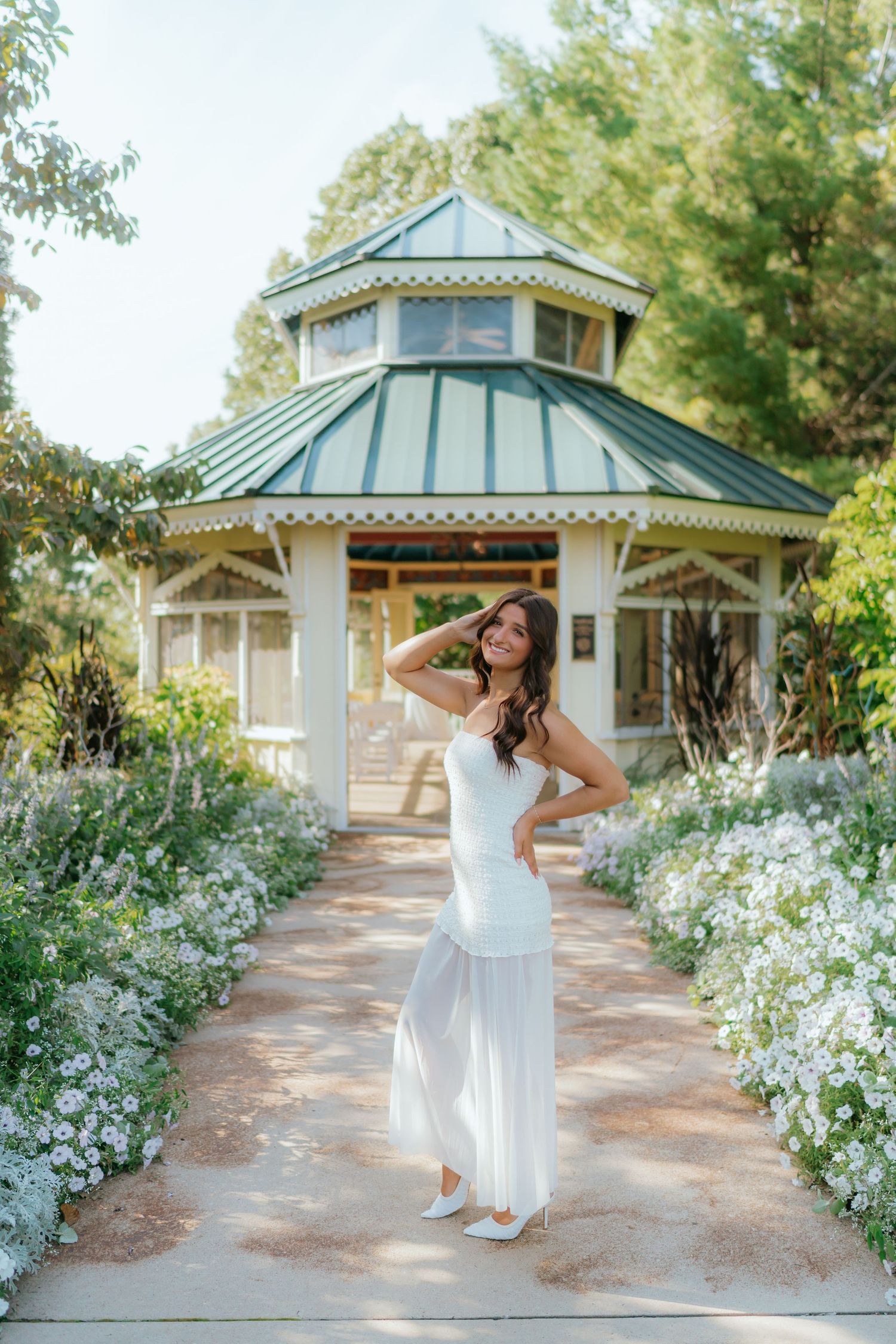 Woman in white dress posing on garden path with gazebo and white flowers in background.