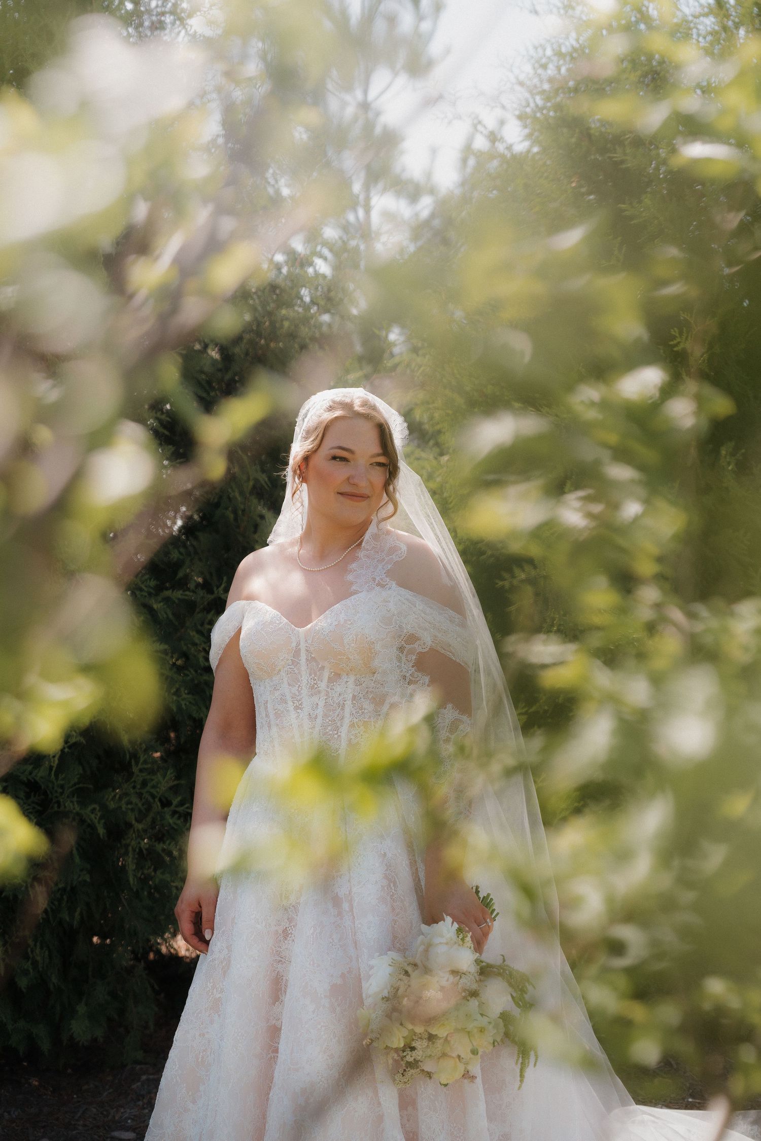 Bride in lace off-shoulder gown and veil holds white bouquet, surrounded by lush greenery in soft sunlight.