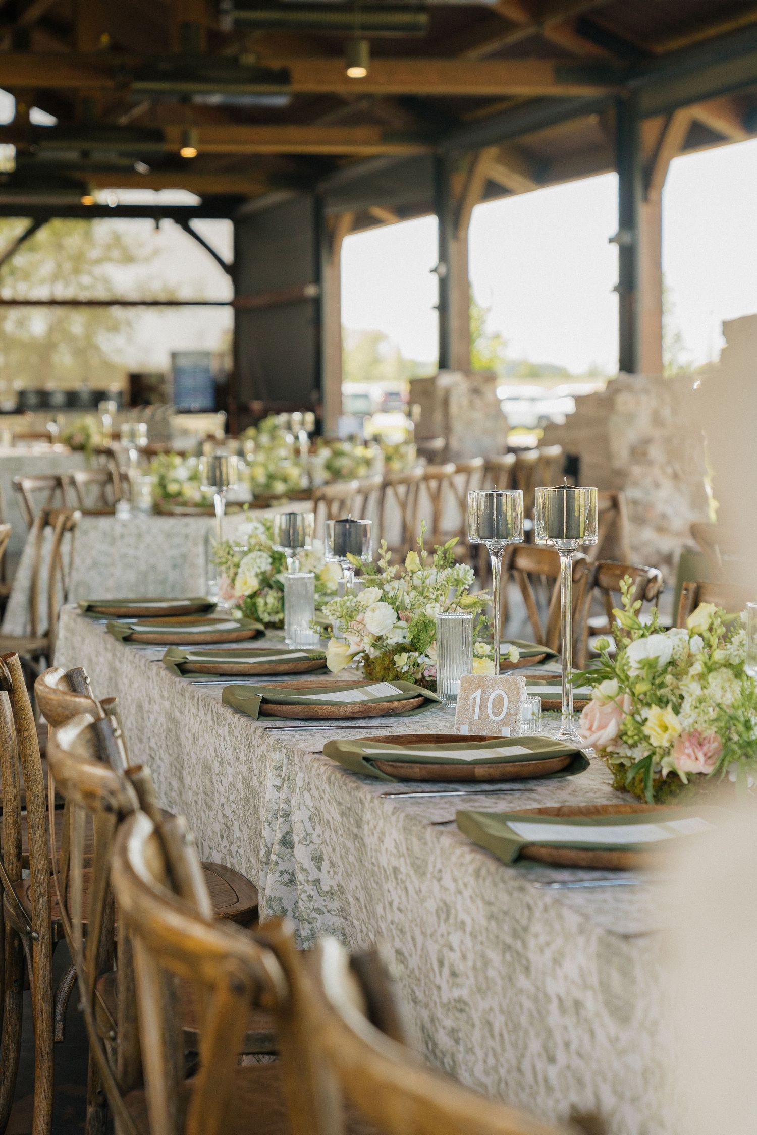 Elegant rustic wedding reception table with floral centerpieces, wooden cross-back chairs, and lace tablecloth under a pavilion.