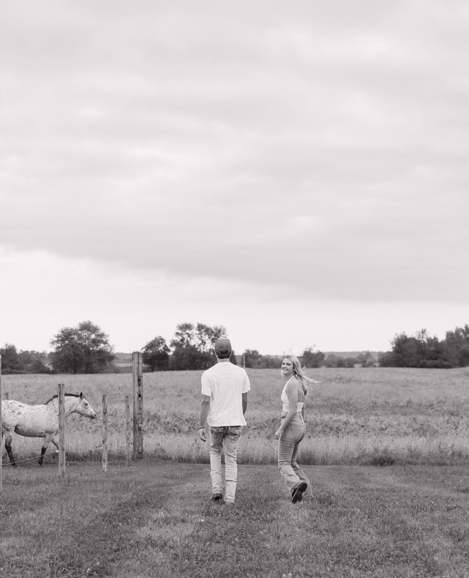 A black and white image shows two people walking in a grassy field with a fence and trees in the background.