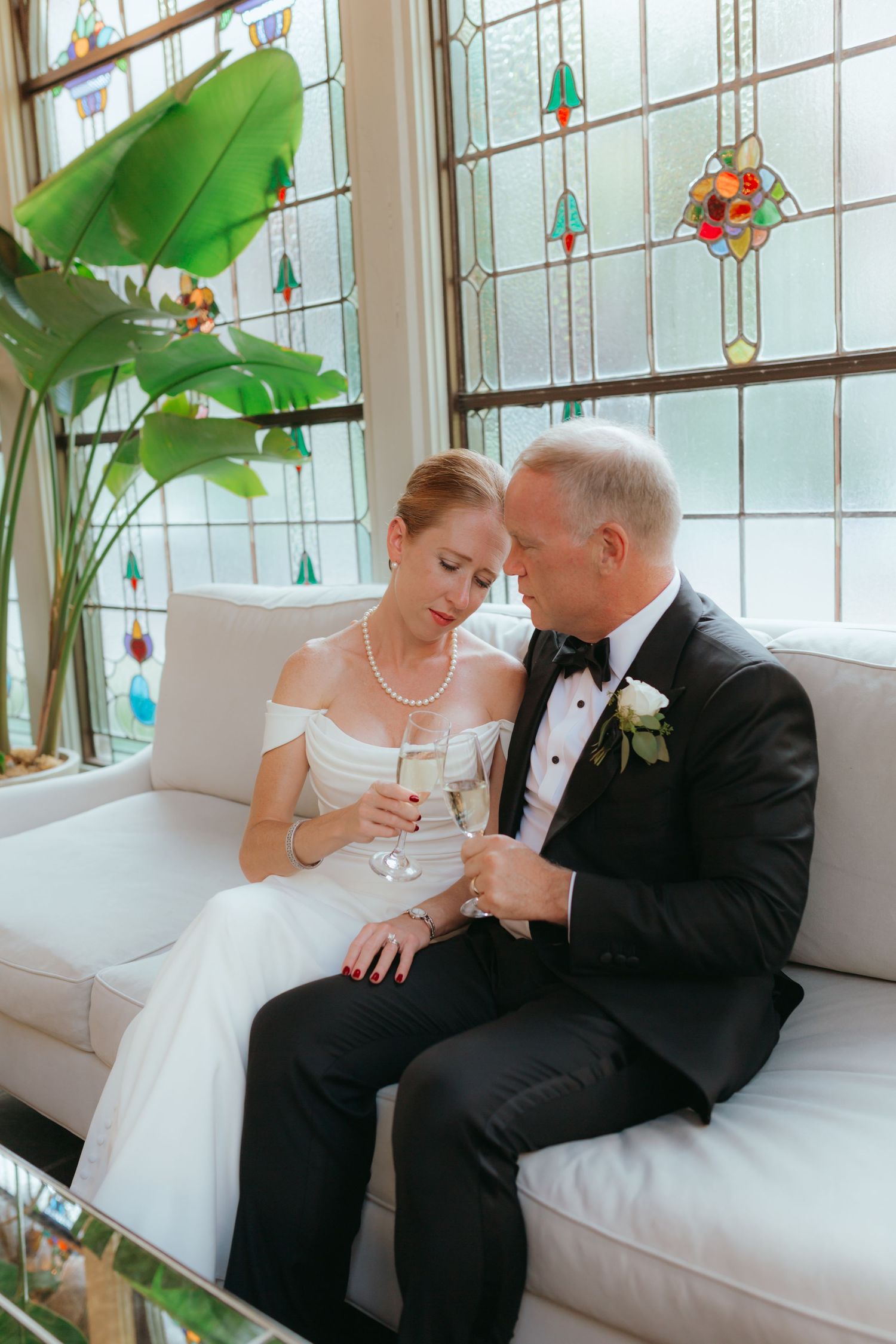 Bride and groom sit together on white couch holding champagne glasses near stained glass windows.