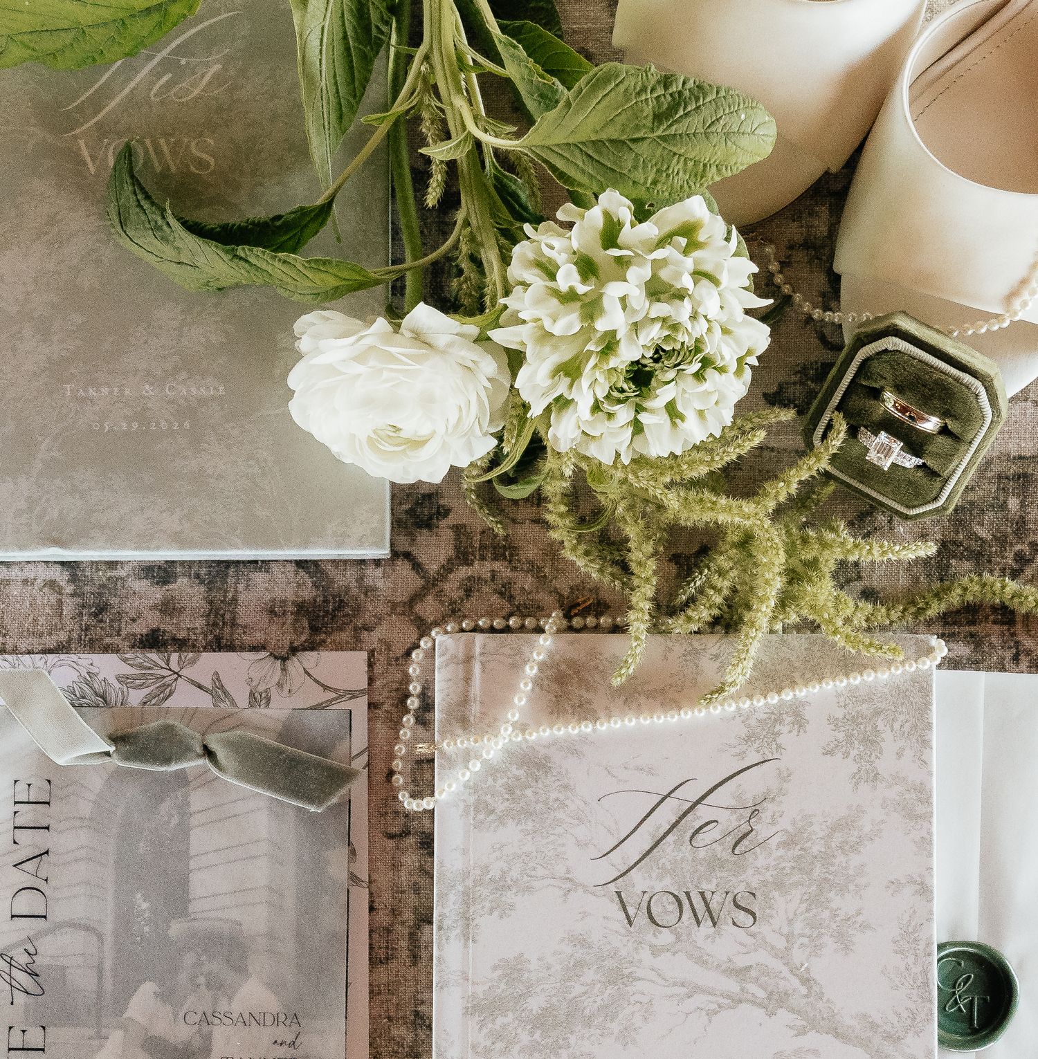 Elegant wedding flat lay with white flowers, vow book, rings, save the date card, and nude heels on a vintage background.