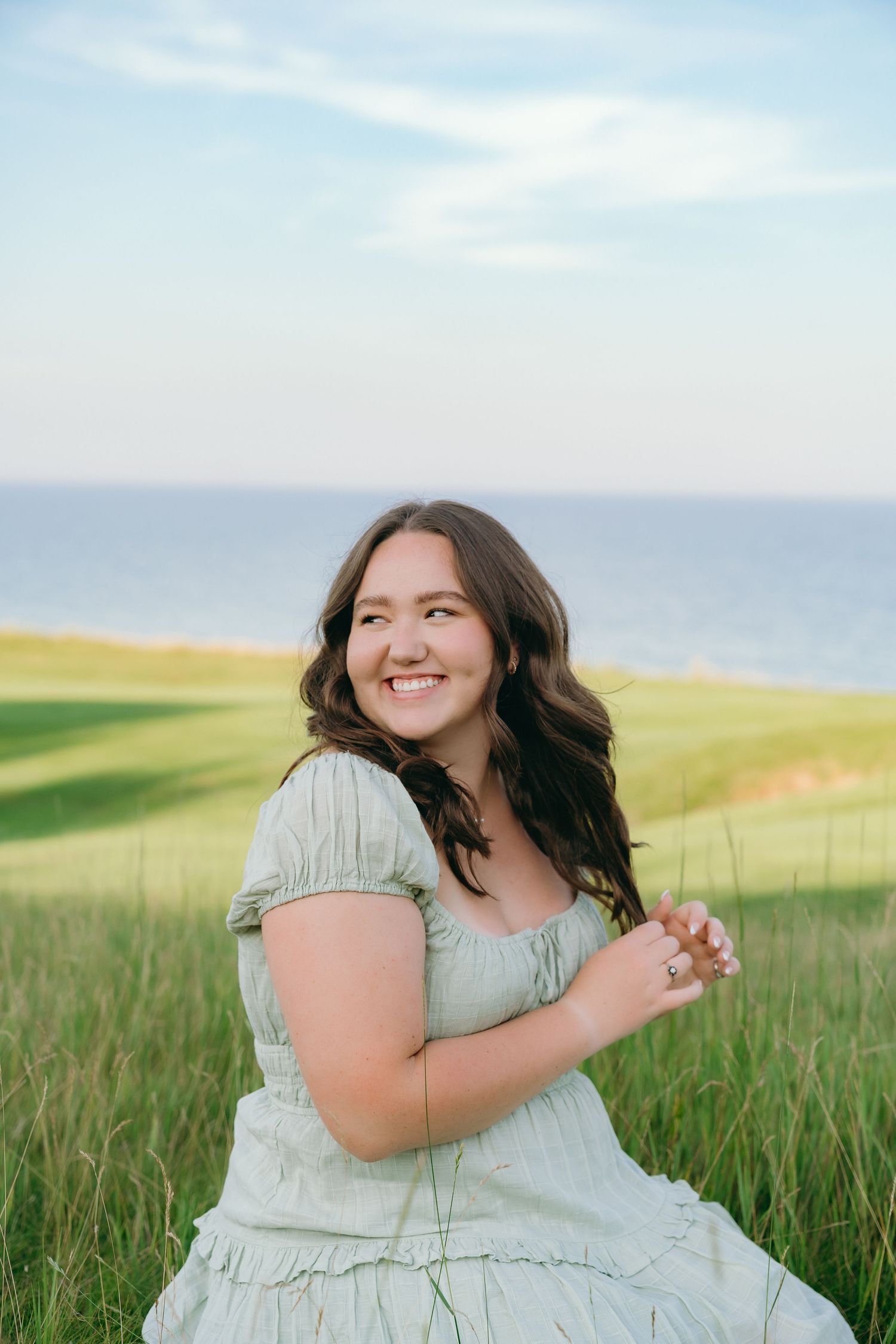 A smiling woman in a light gray dress stands in a grassy field with a scenic ocean view in the background.