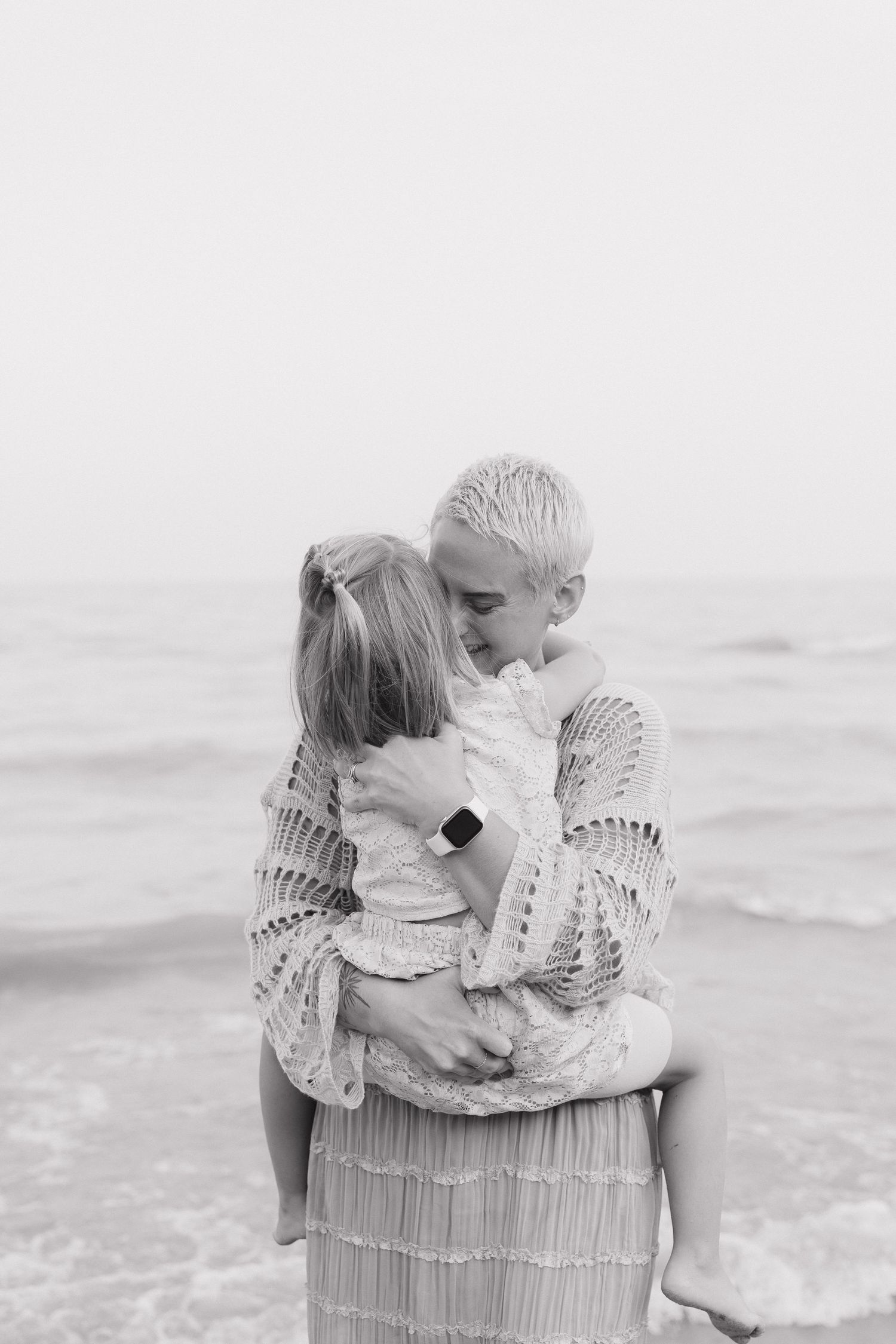 A black and white image shows a mother and daughter on a beach, with the ocean visible in the background.