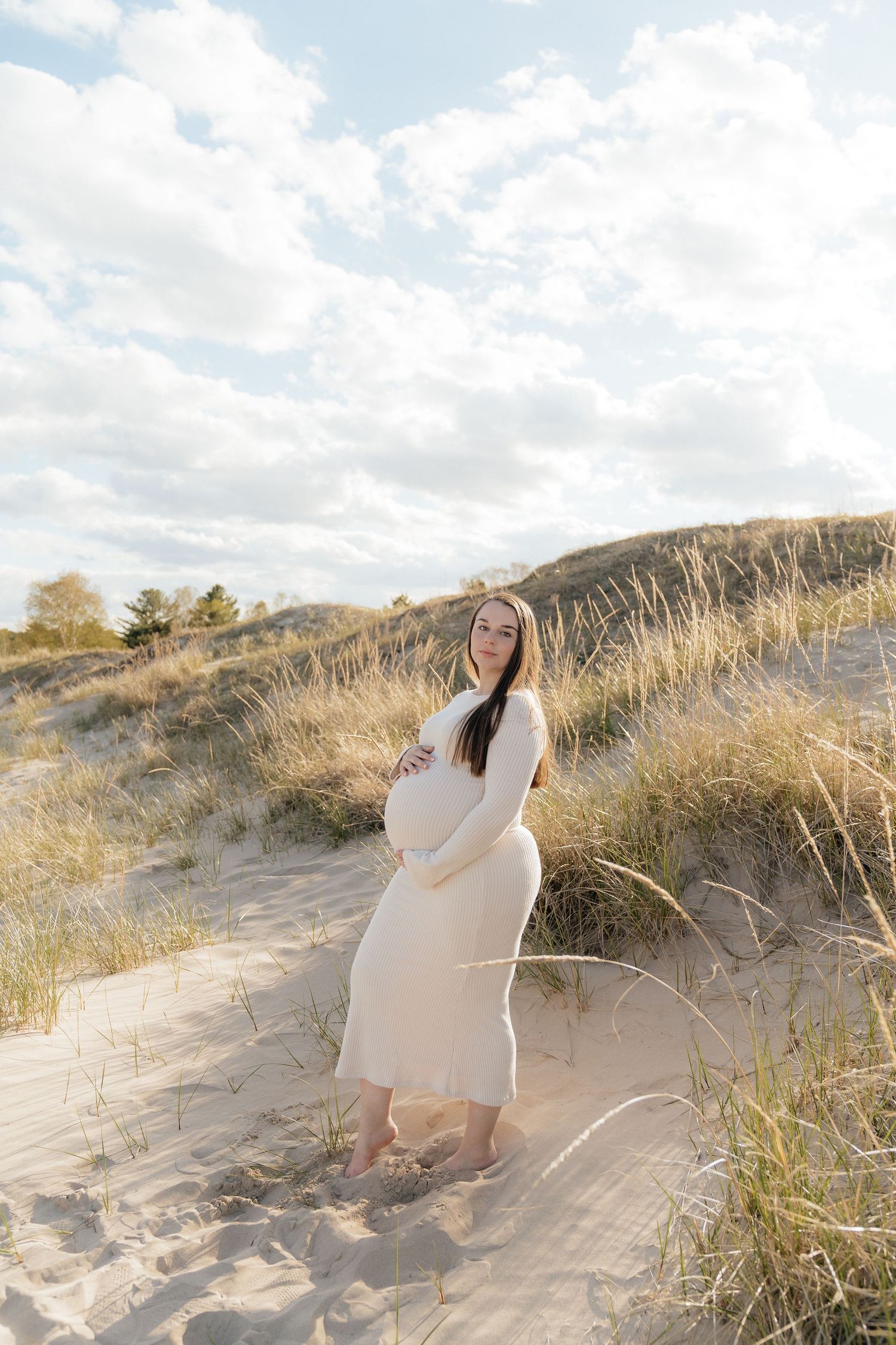 Pregnant woman in white dress cradling baby bump on sandy dunes with golden grass and cloudy sky.