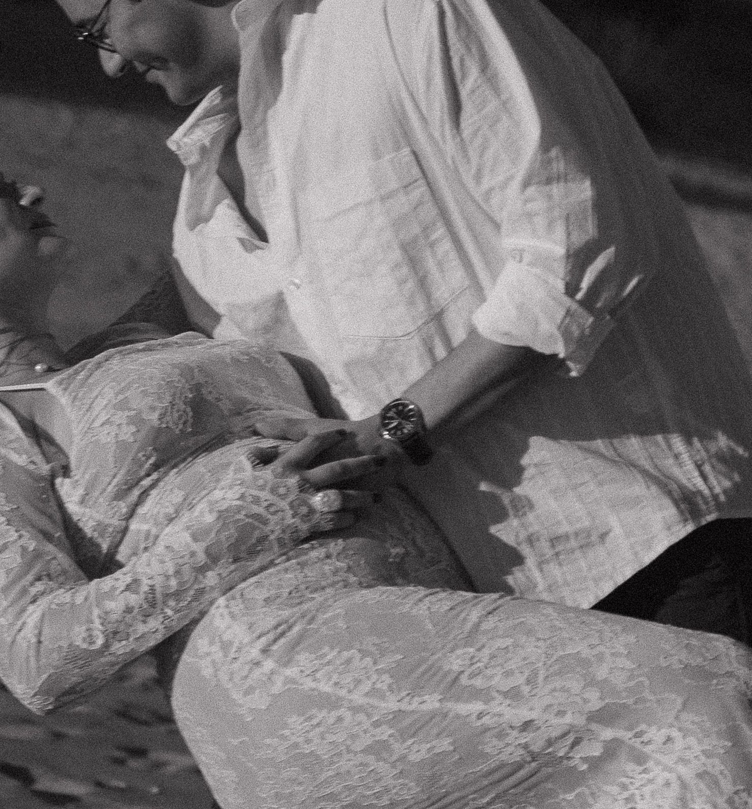 Black and white photo of two people in white lace and linen outfits sitting closely together outdoors.