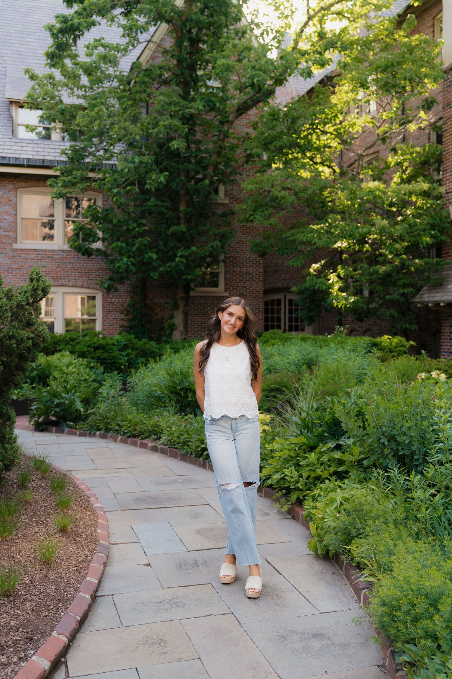 Young woman in white top and ripped jeans smiling on a stone path surrounded by lush gardens near a brick building.