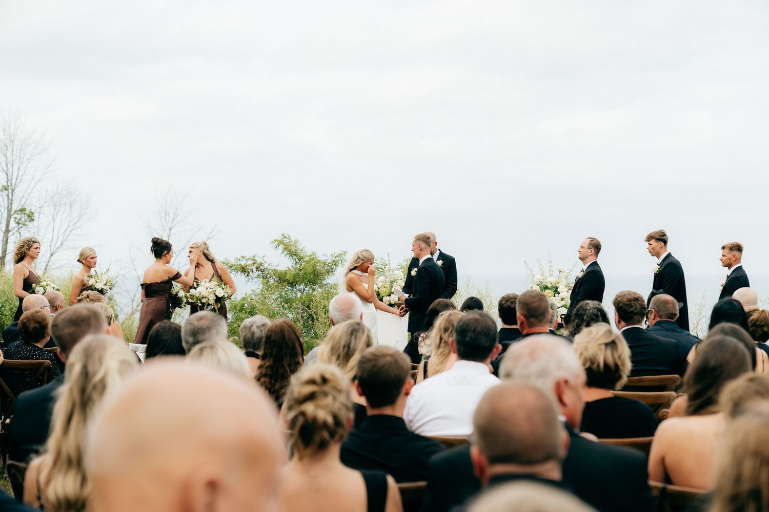 Outdoor wedding ceremony with bride and groom at altar, wedding party standing alongside, guests seated in rows under cloudy sky.