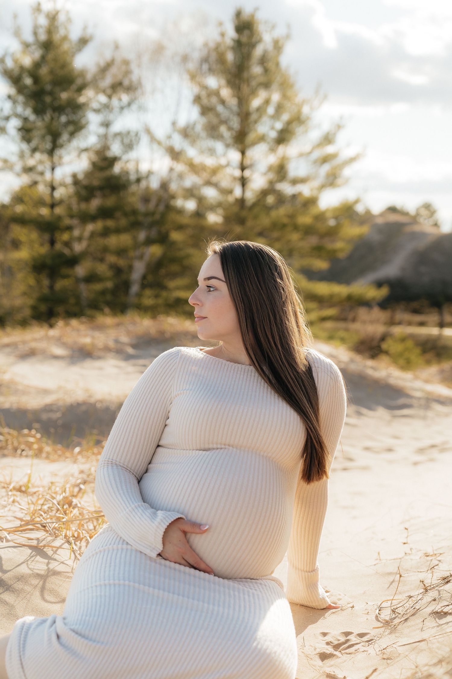 Pregnant woman in cream ribbed dress cradling baby bump on sandy dunes with pine trees in golden hour light.