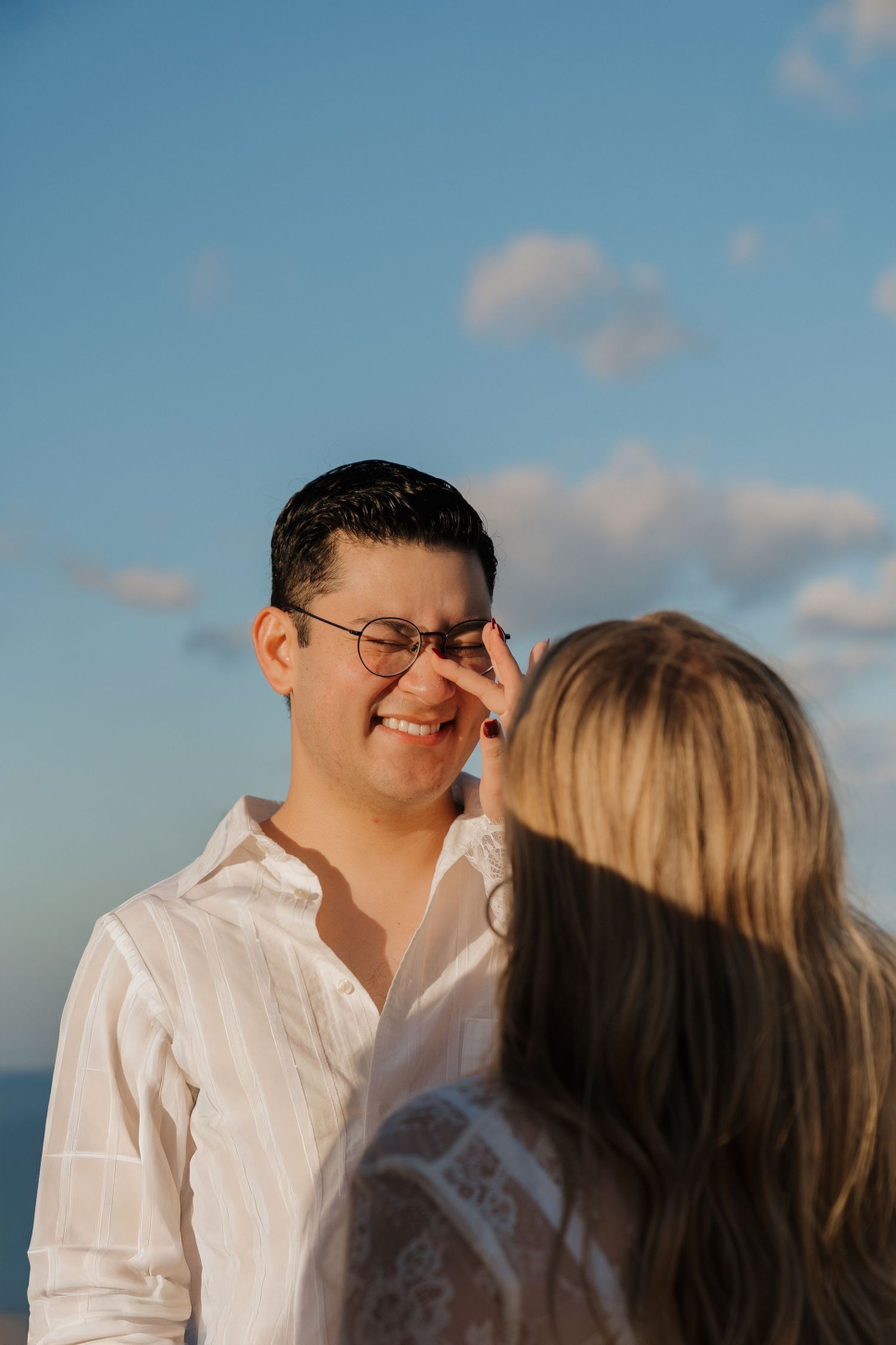 Smiling man with glasses in white shirt looks at blonde woman during golden hour beach photo session.