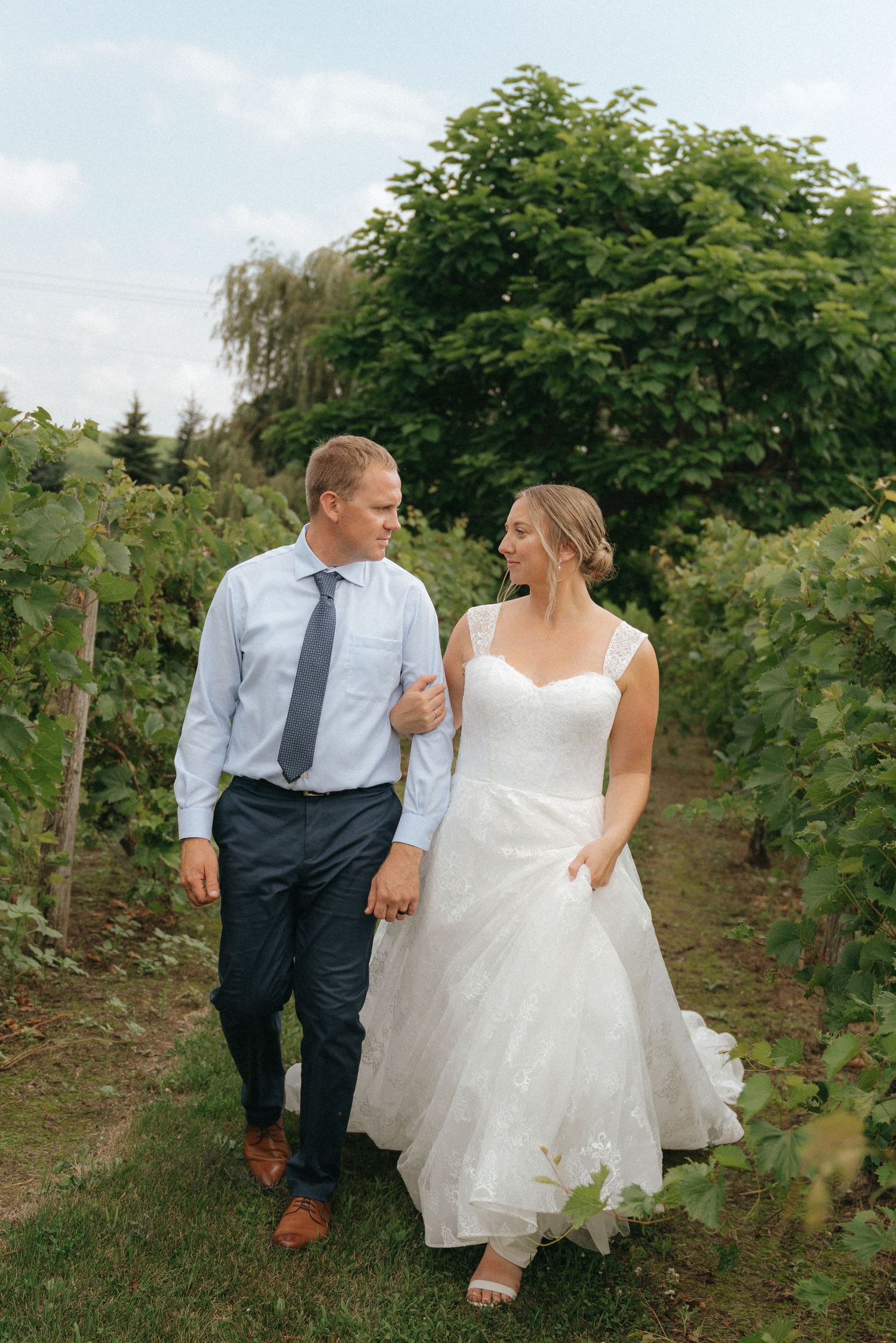 A bride and groom walk hand-in-hand through a lush, green vineyard on their wedding day.