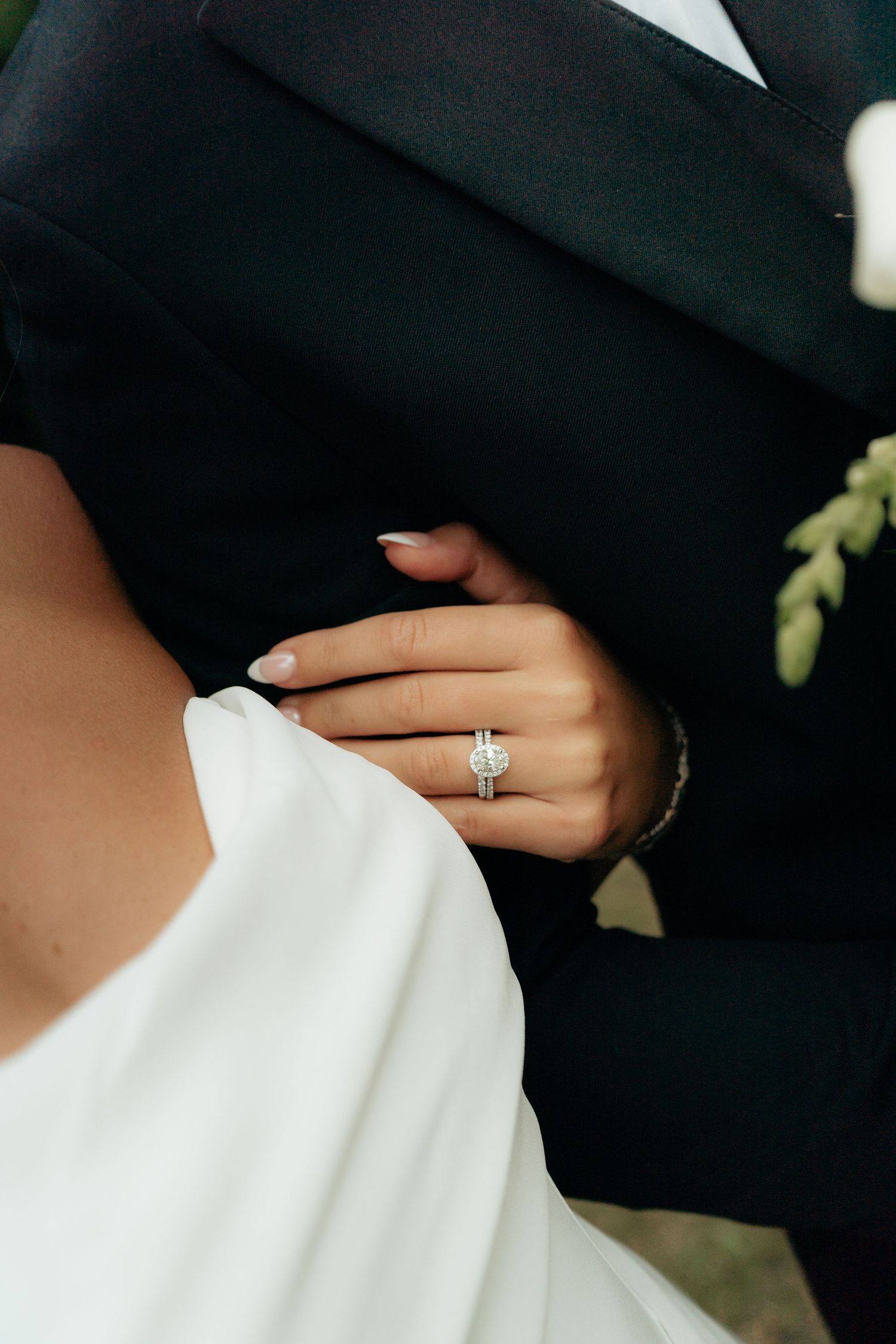 Bride showcasing a stunning diamond halo engagement ring while holding grooms arm in black suit on wedding day.