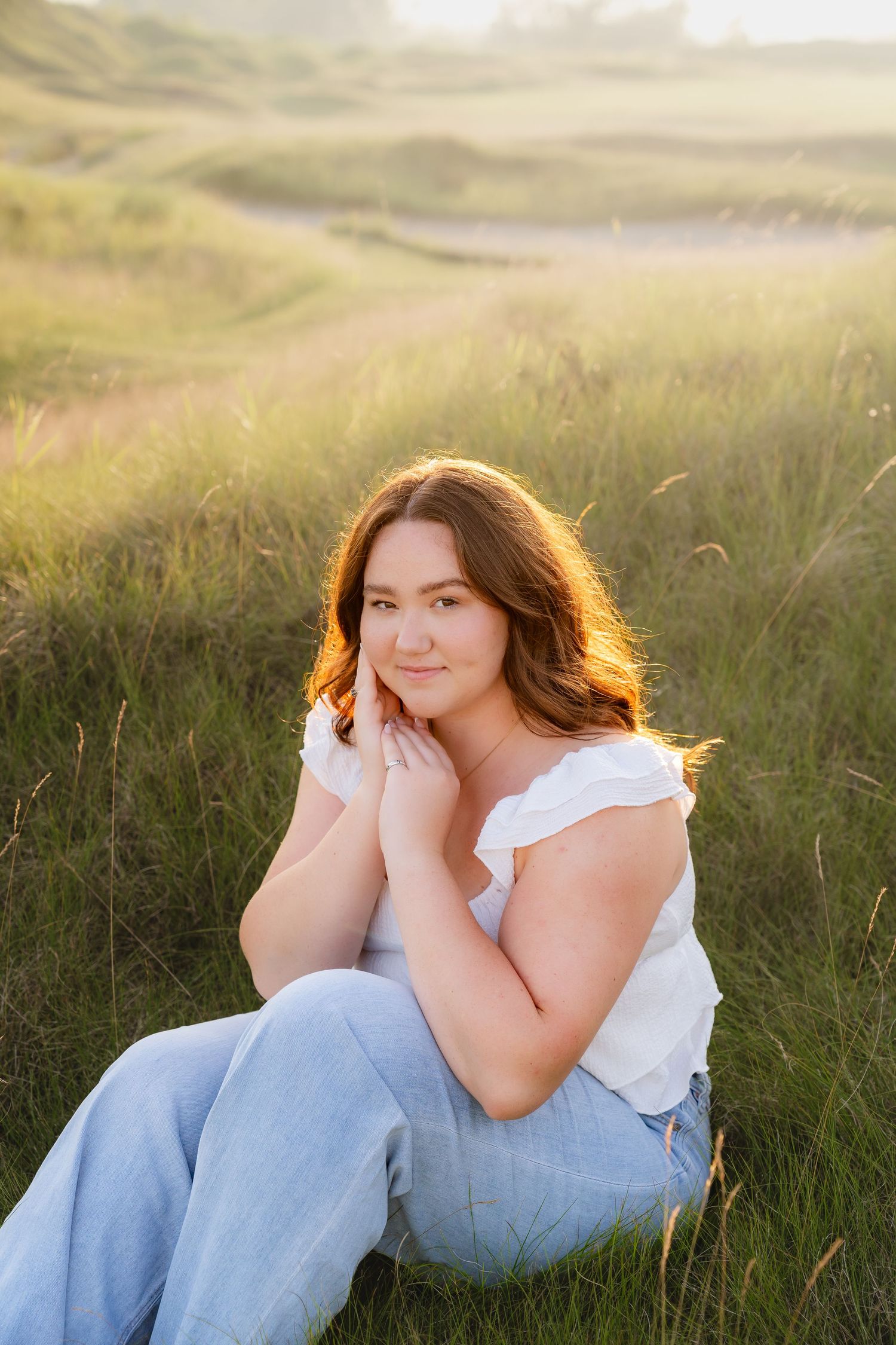 A young woman with long, wavy brown hair sits in a grassy field, smiling and looking off to the side.