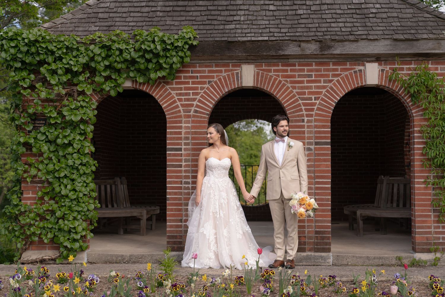 A bride and groom stand together in front of an arched brick building surrounded by lush greenery and flowers.